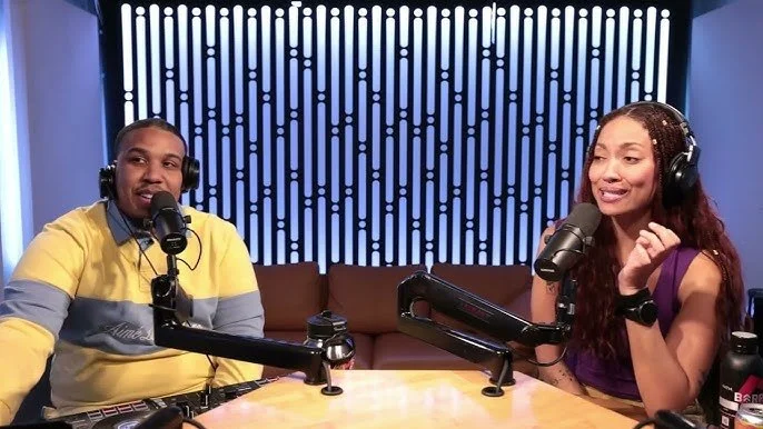 Two people, a man and a woman, are sitting at a table with microphones, recording a podcast or radio show in a studio with a patterned blue background.