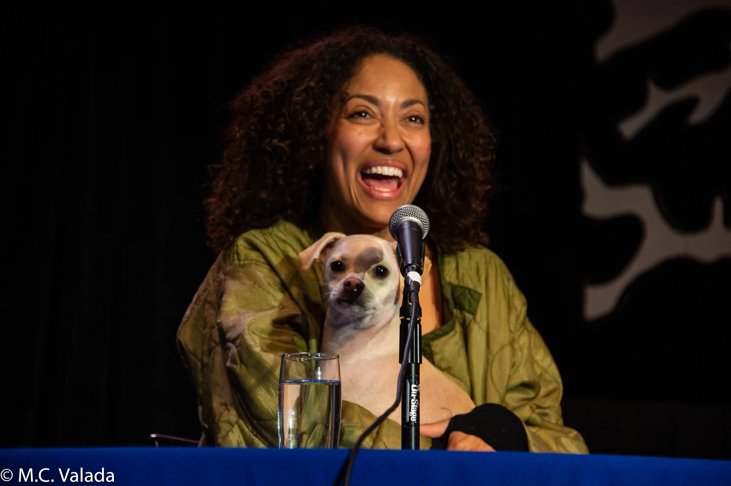 A woman with curly hair, wearing a green jacket, laughing while holding a small tan dog at a table with a microphone and a glass of water. The background is black.