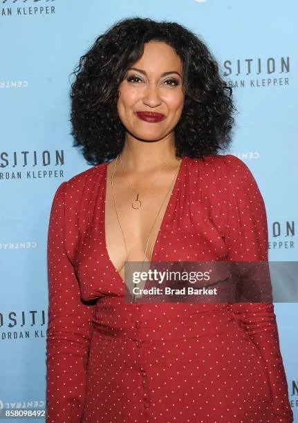 Woman with curly black hair wearing a red polka-dot dress and a gold necklace, standing against a blue backdrop with text.