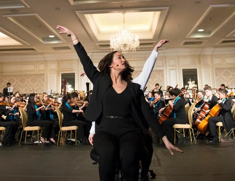A woman leading a dance or theatrical performance in front of an orchestra in a grand, beautifully decorated concert hall with a chandelier.