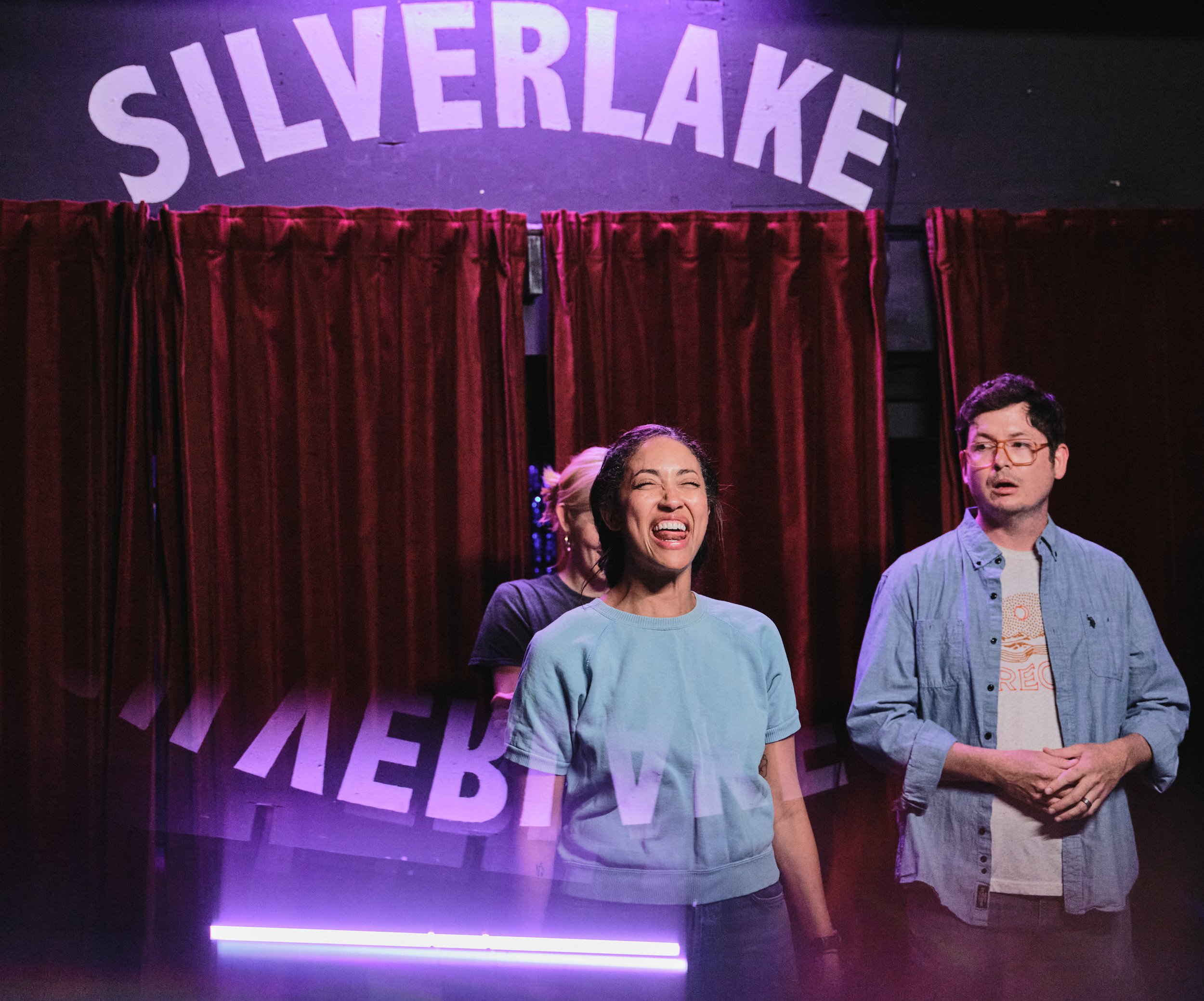 A woman smiling and a man standing next to each other in front of a red curtain at Silverlake. The woman is wearing a pastel blue T-shirt and the man is wearing a light denim shirt over a T-shirt. The setting appears to be a comedy or entertainment v