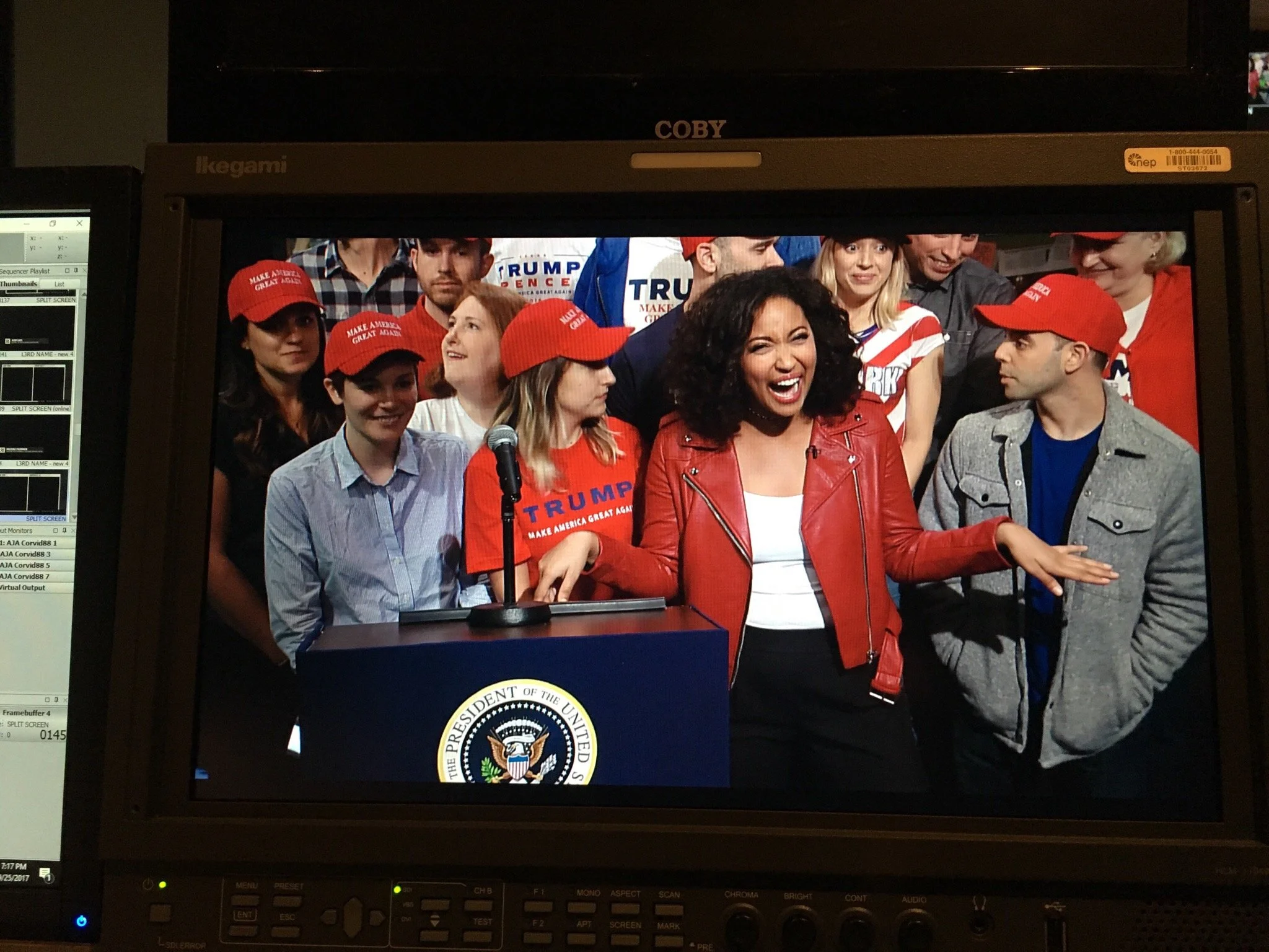 A woman in a red leather jacket laughing and gesturing with her hand at a political rally with supporters wearing "Make America Great Again" hats and shirts, standing behind a podium with the presidential seal.