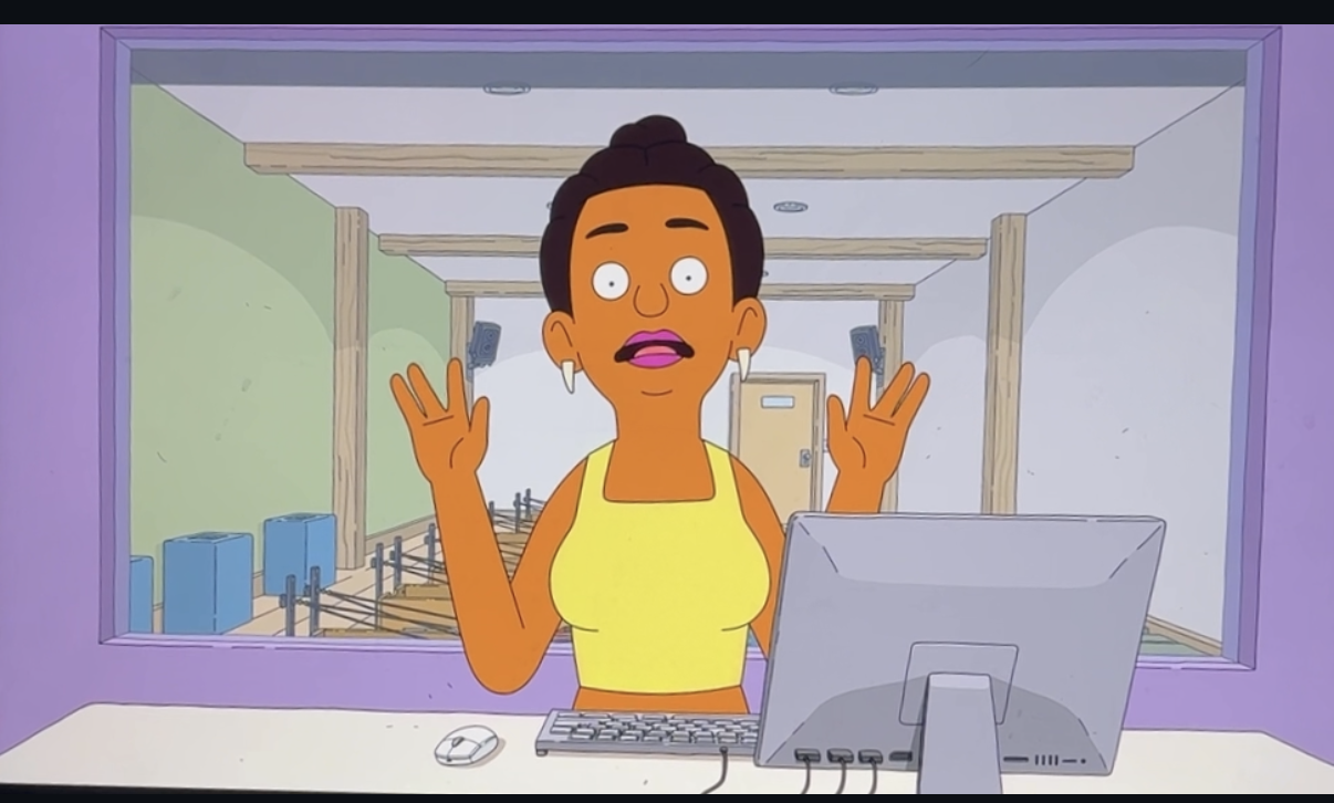 Animated woman with dark hair in a bun, wearing a yellow top, appears surprised and is gesturing with her hands while talking in front of a computer, through a glass window that shows a gym with wooden beams and exercise equipment.