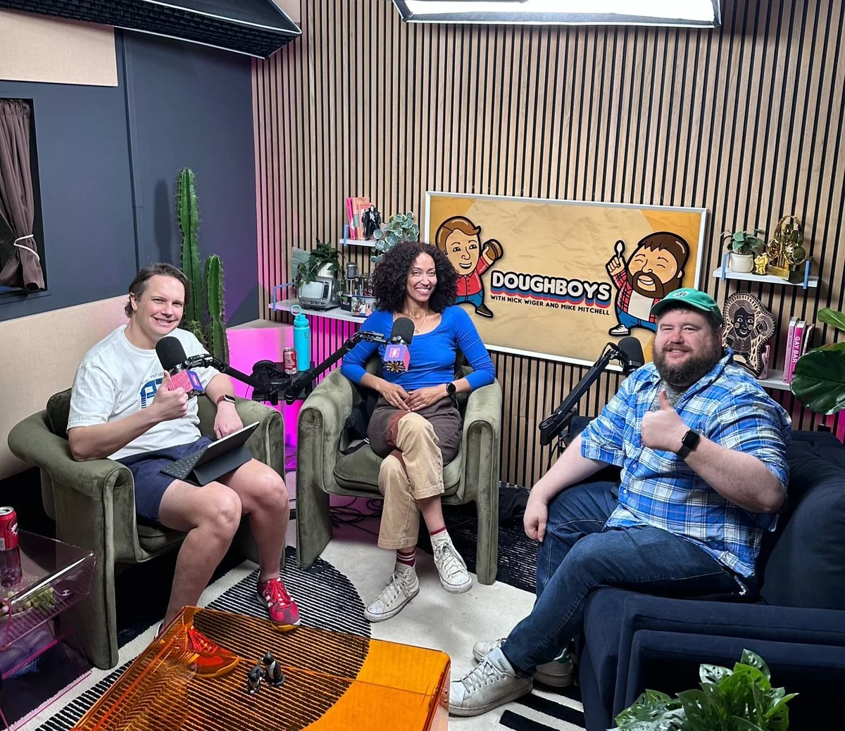 Three people sit in a recording studio for the Doughboys podcast, smiling at the camera. A woman in the center is seated in an armchair, flanked by two men in armchairs. Behind them is a sign for the Doughboys podcast, and the room has various decora