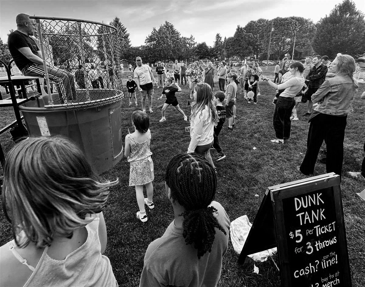 People gathered at an outdoor fair playing a dunk tank game with a boy sitting on the dunk tank's seat. A sign nearby reads: 'Dunk Tank $5 per ticket for 3 throws. Join the line!'