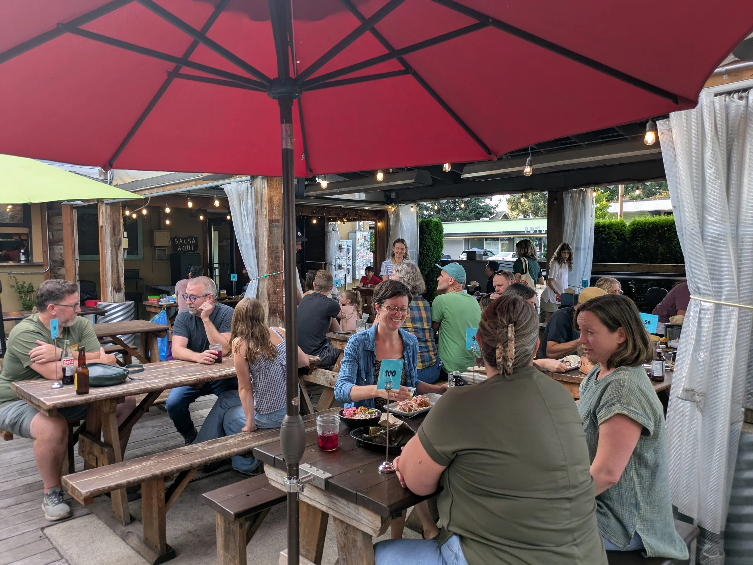 People dining at outdoor wooden tables under large umbrellas, with string lights hanging above, at a busy restaurant or café.