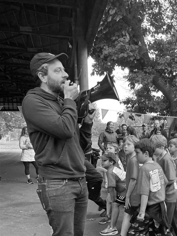 A man with a beard and cap speaking into a megaphone at an outdoor event, with children and adults standing behind him under a pavilion.