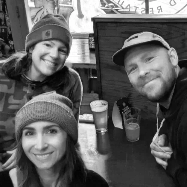 Three people smiling at a table with drinks in a casual setting, possibly a bar or restaurant.