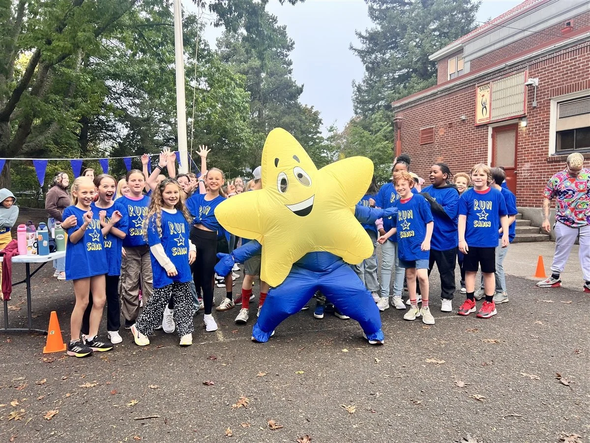 A group of student runners in matching blue t-shirts posing with the school macot, a yellow star with a smiley face on it.
