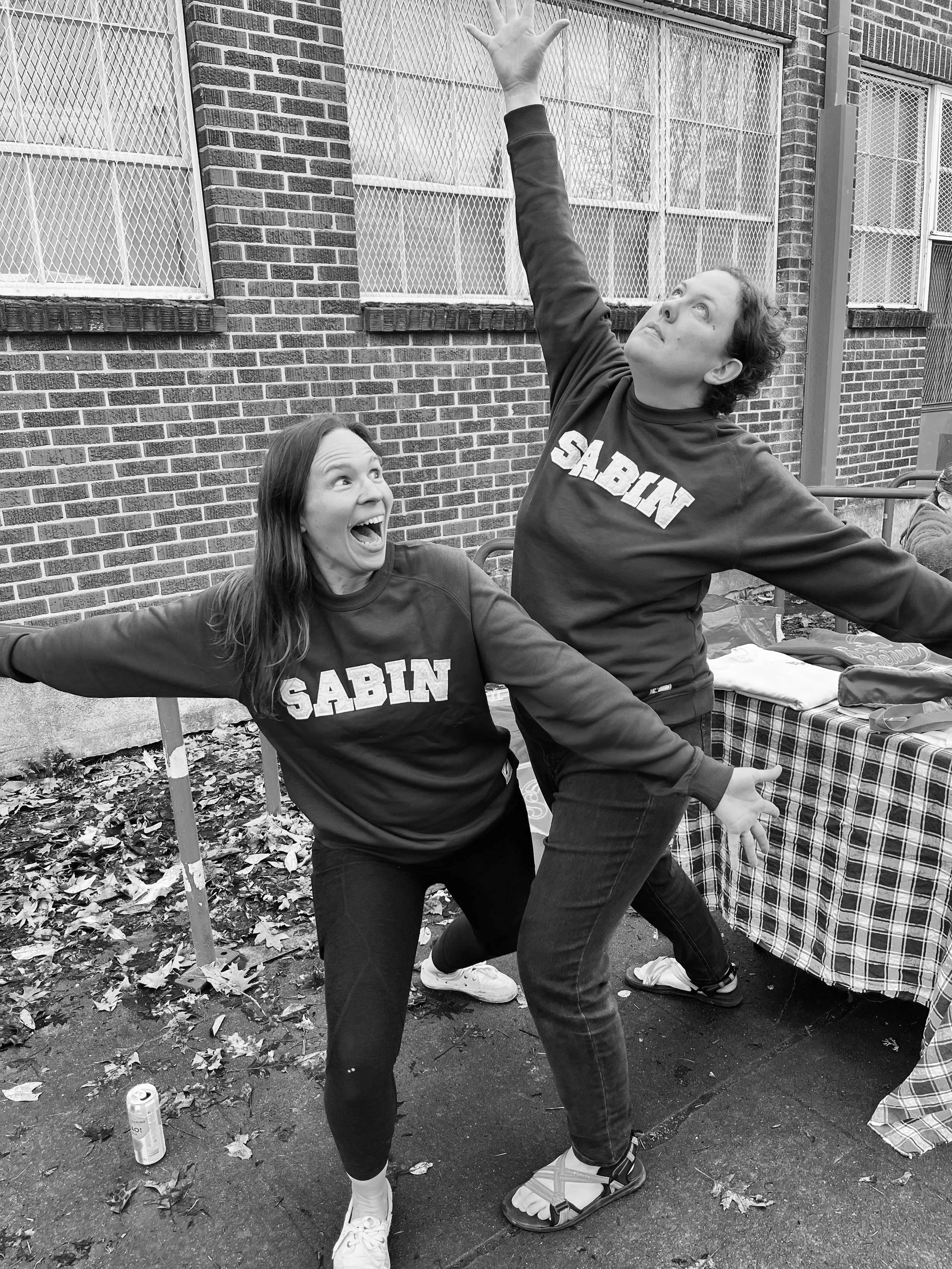 Two women wearing matching sweatshirts with the name 'SABIN' are posing playfully outdoors, with one woman reaching upwards and the other with arms outstretched.
