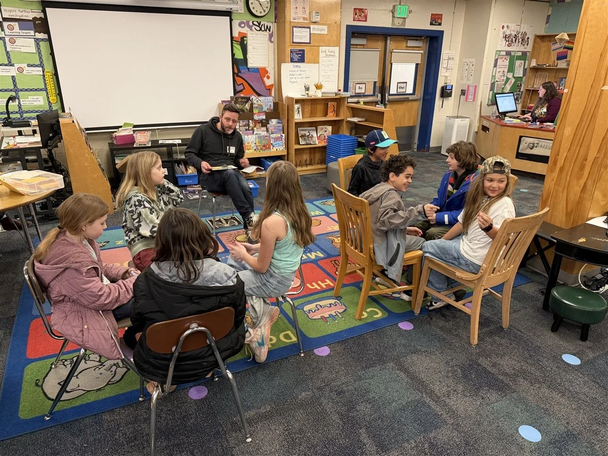 Group of children sitting in small groups in a classroom, engaging in conversations, with a teacher or instructor nearby and a teacher working at a computer in the background.