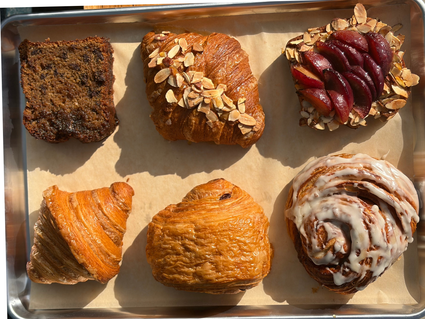 Six assorted pastries on a metal tray, including a rectangular chocolate chip bread, a croissant with sliced almonds, a berry-topped pastry with almonds, a plain croissant, a Danish with a fruit filling, and a cinnamon roll with white icing.