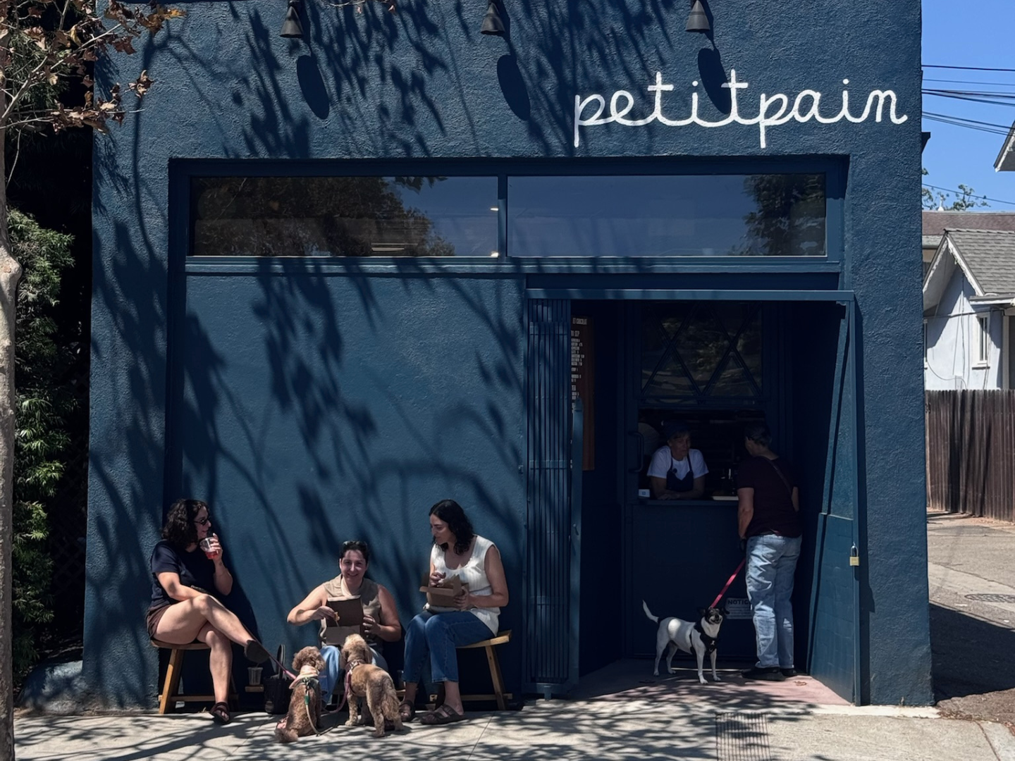 People sitting outside Petitpain cafe with dogs, two customers at the counter, and the blue facade of the cafe.
