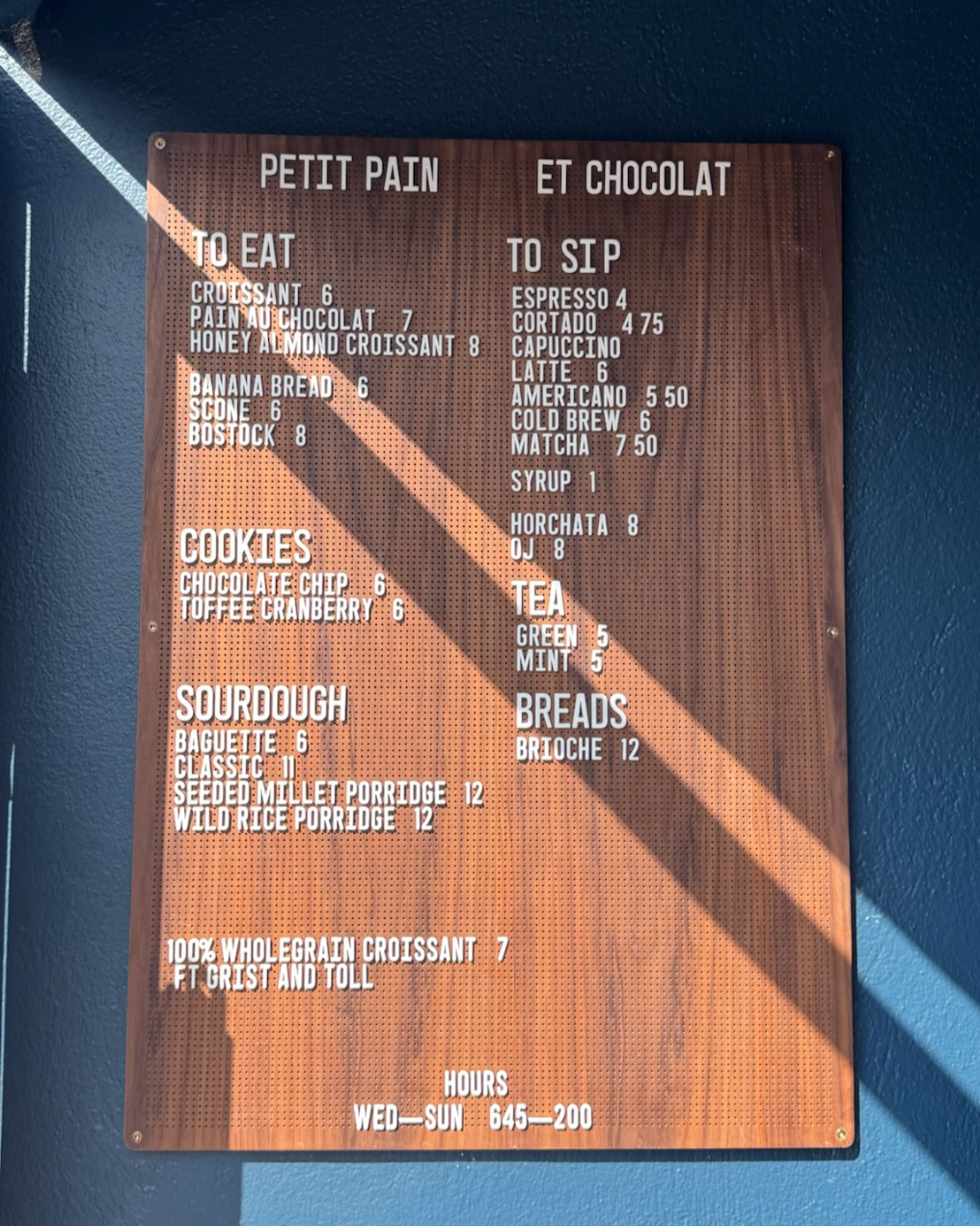 Wooden menu board with French section titles, listing various baked goods, cookies, sourdough, bread, and beverages such as coffee and tea, with prices.