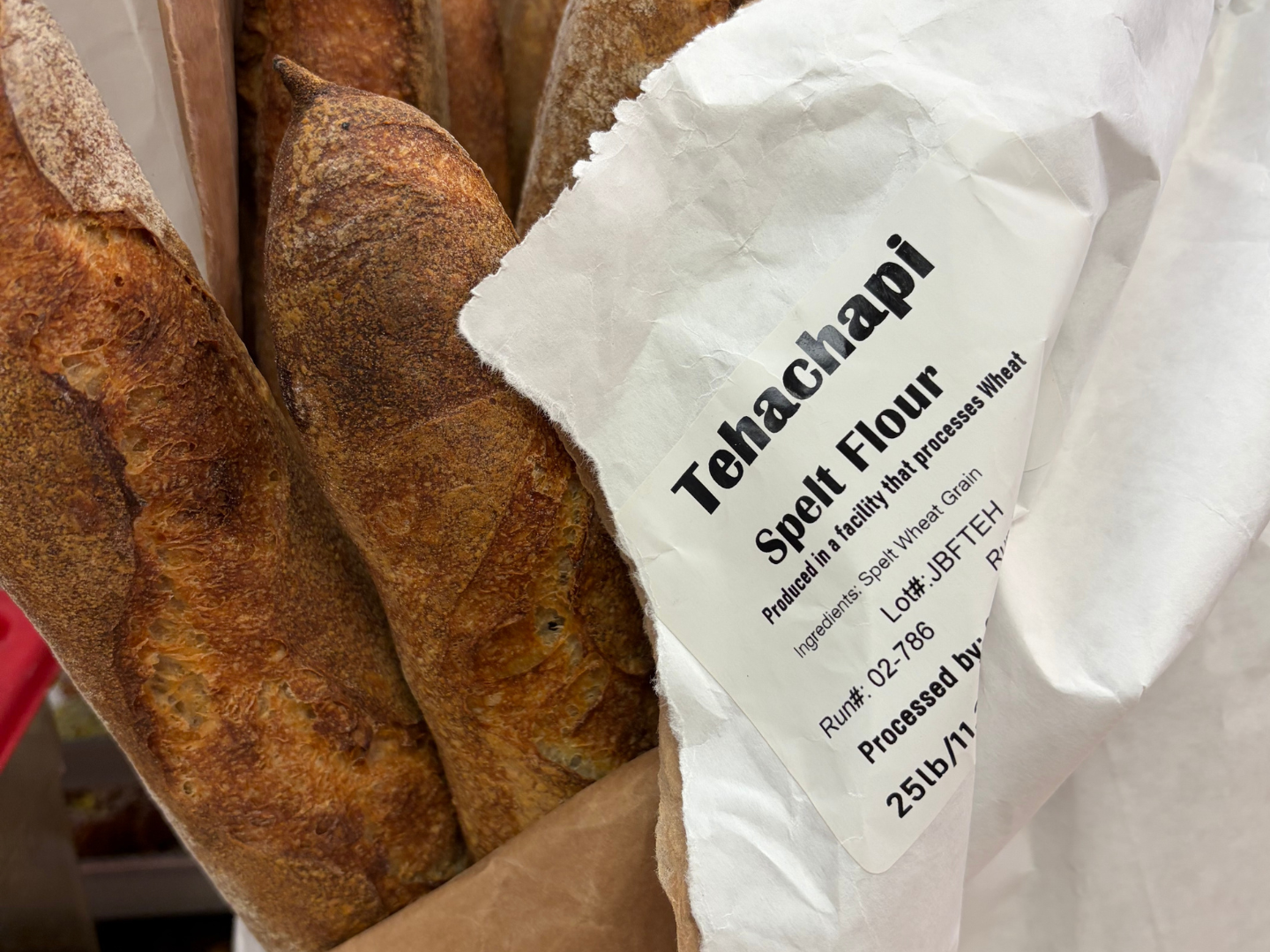 Two baguettes of toasted bread in paper packaging labeled 'Tehachaapi Spelt Flour,' produced in a facility that processes wheat, with a weight of 25 pounds.