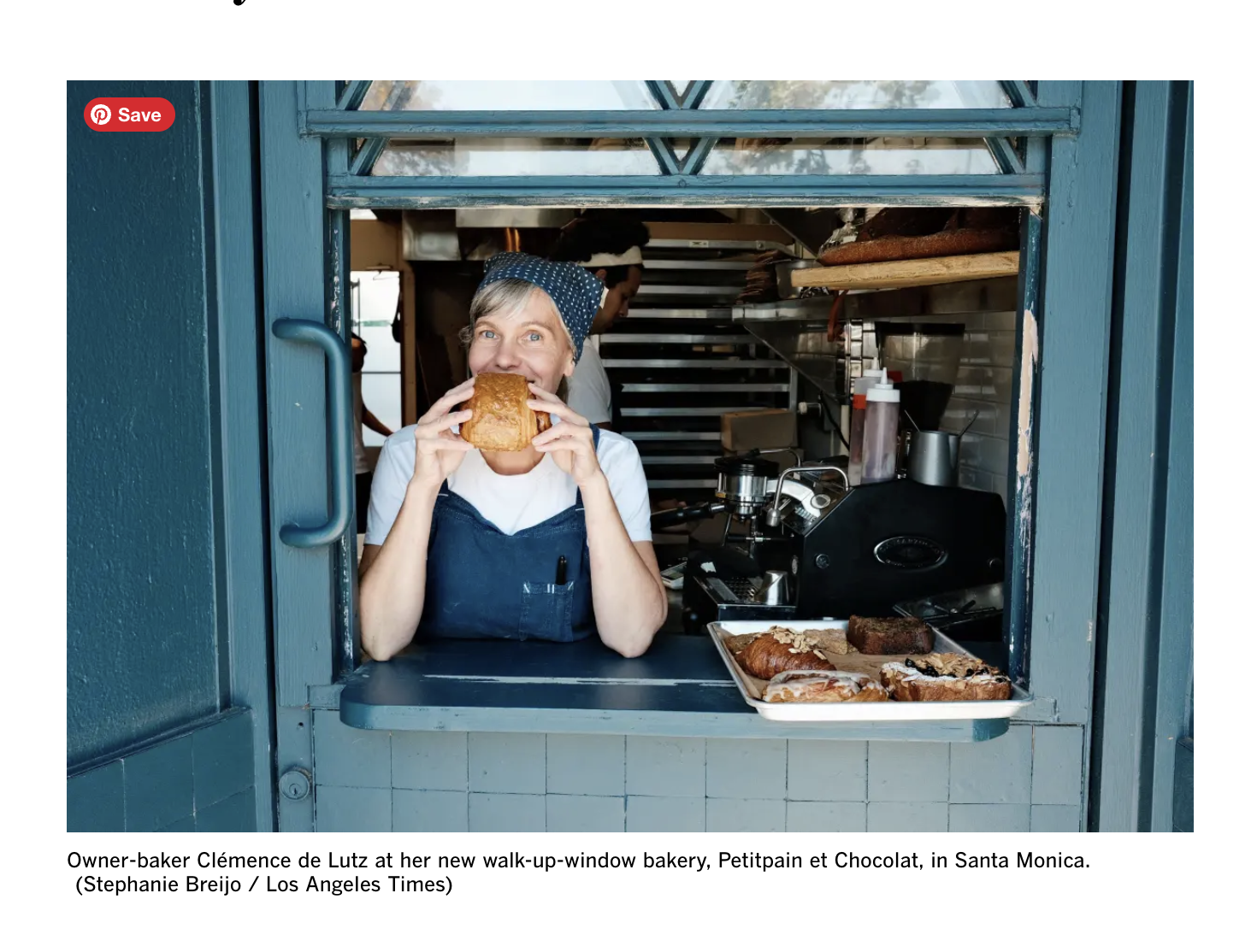 Owner-baker Clémence de Lutz in her walk-up-window bakery, Petitpain et Chocolat, in Santa Monica, smiling and biting into a piece of bread with baked goods on a tray in front of her.