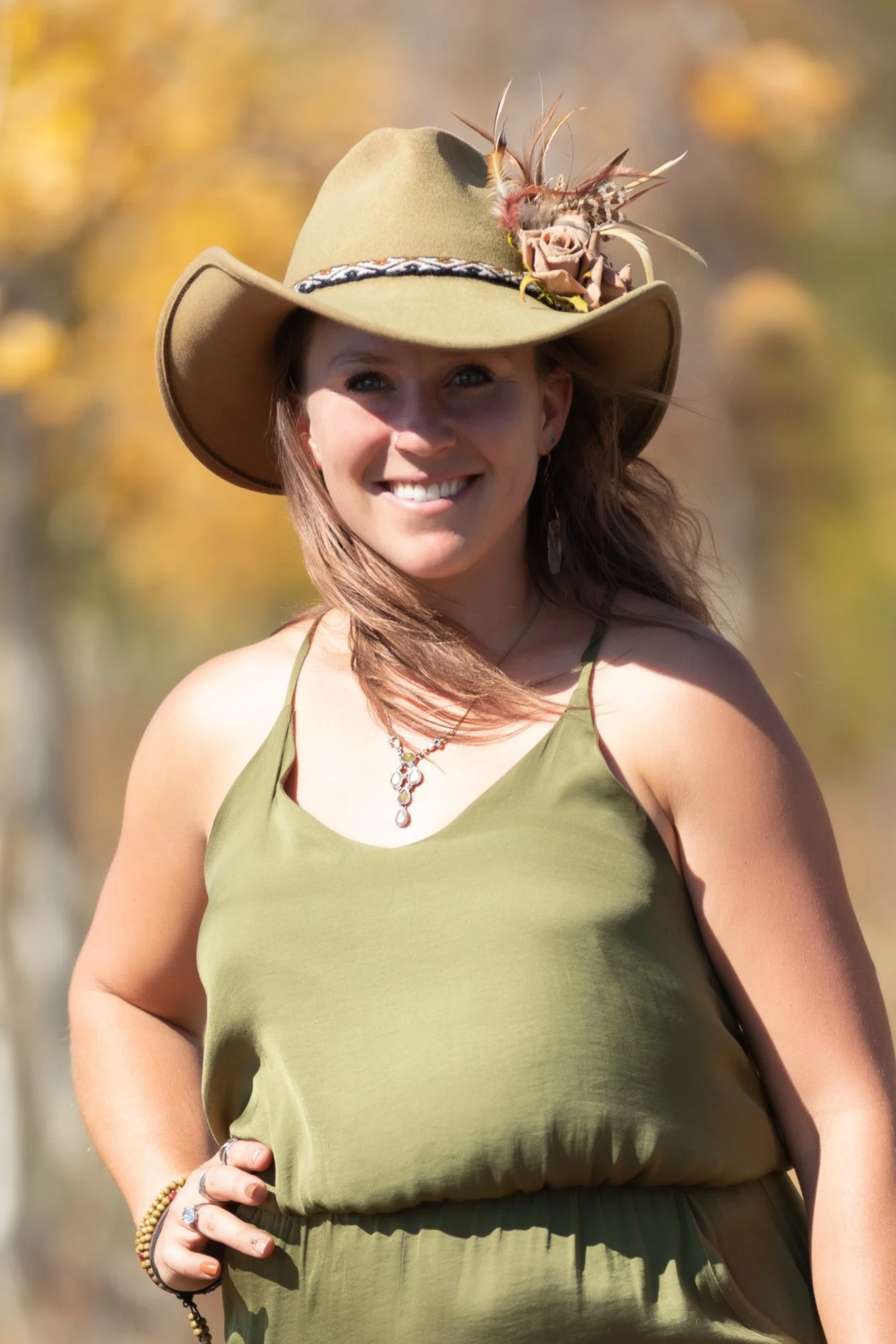 A woman smiling outdoors wearing a green dress and a tan wide-brim hat decorated with flowers and feathers.