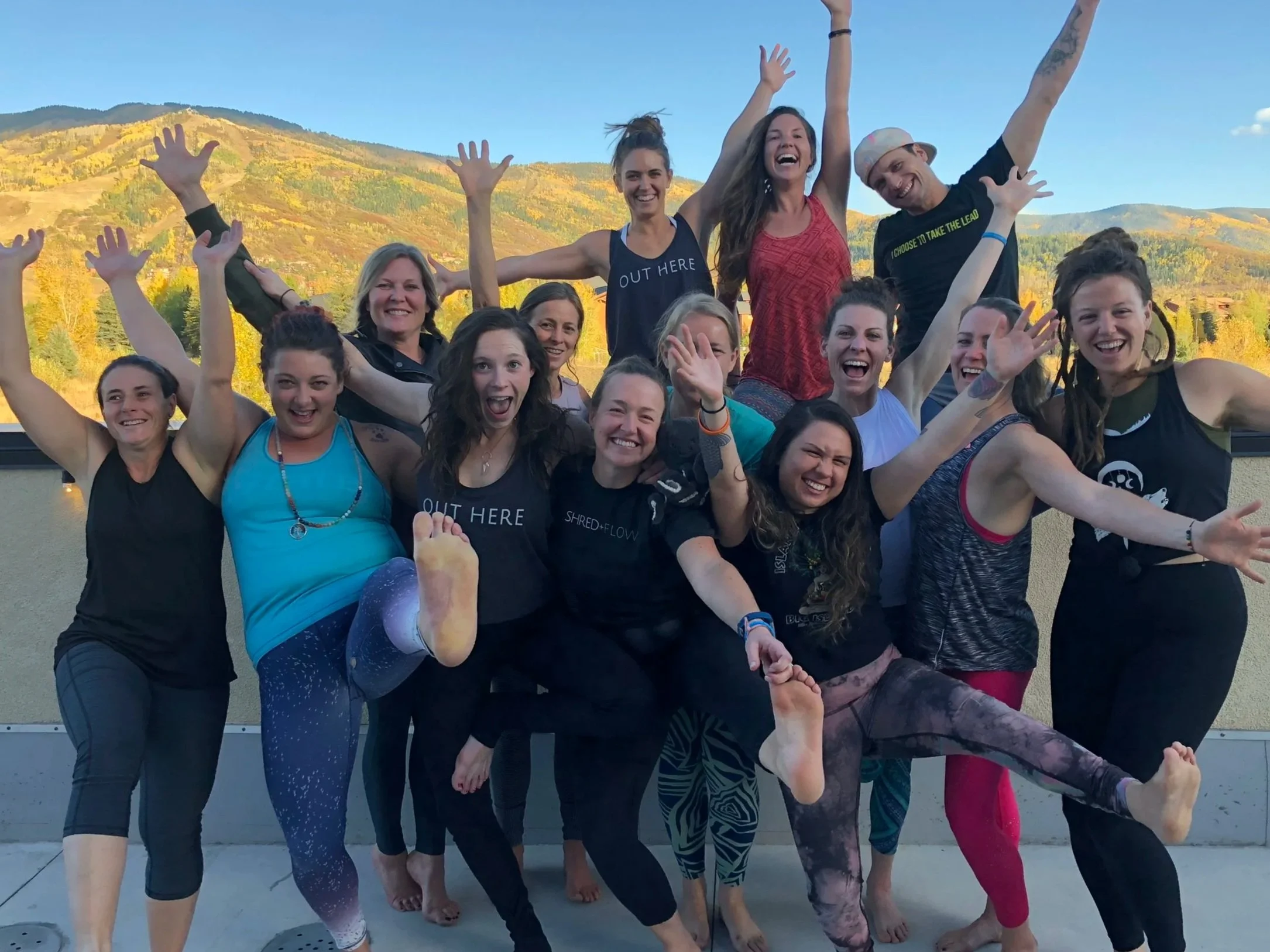 Group of smiling women practicing yoga outdoors on a rooftop with mountain scenery in the background, some standing on one leg, in balance poses, wearing colorful activewear.