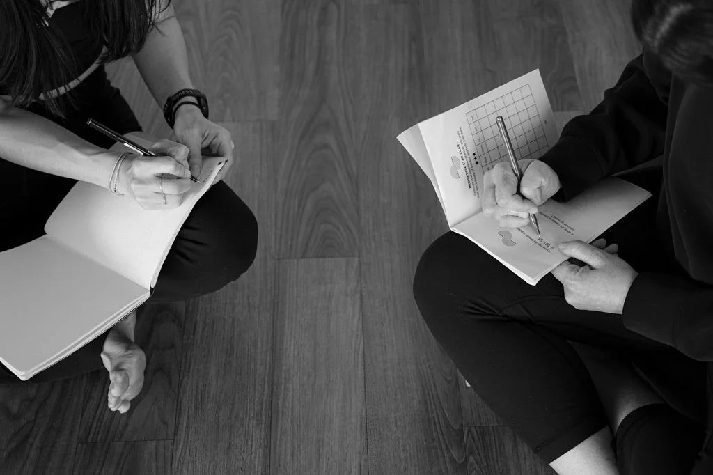 Two people sitting on a wooden floor, writing in notebooks, with papers and pens, viewed from above.