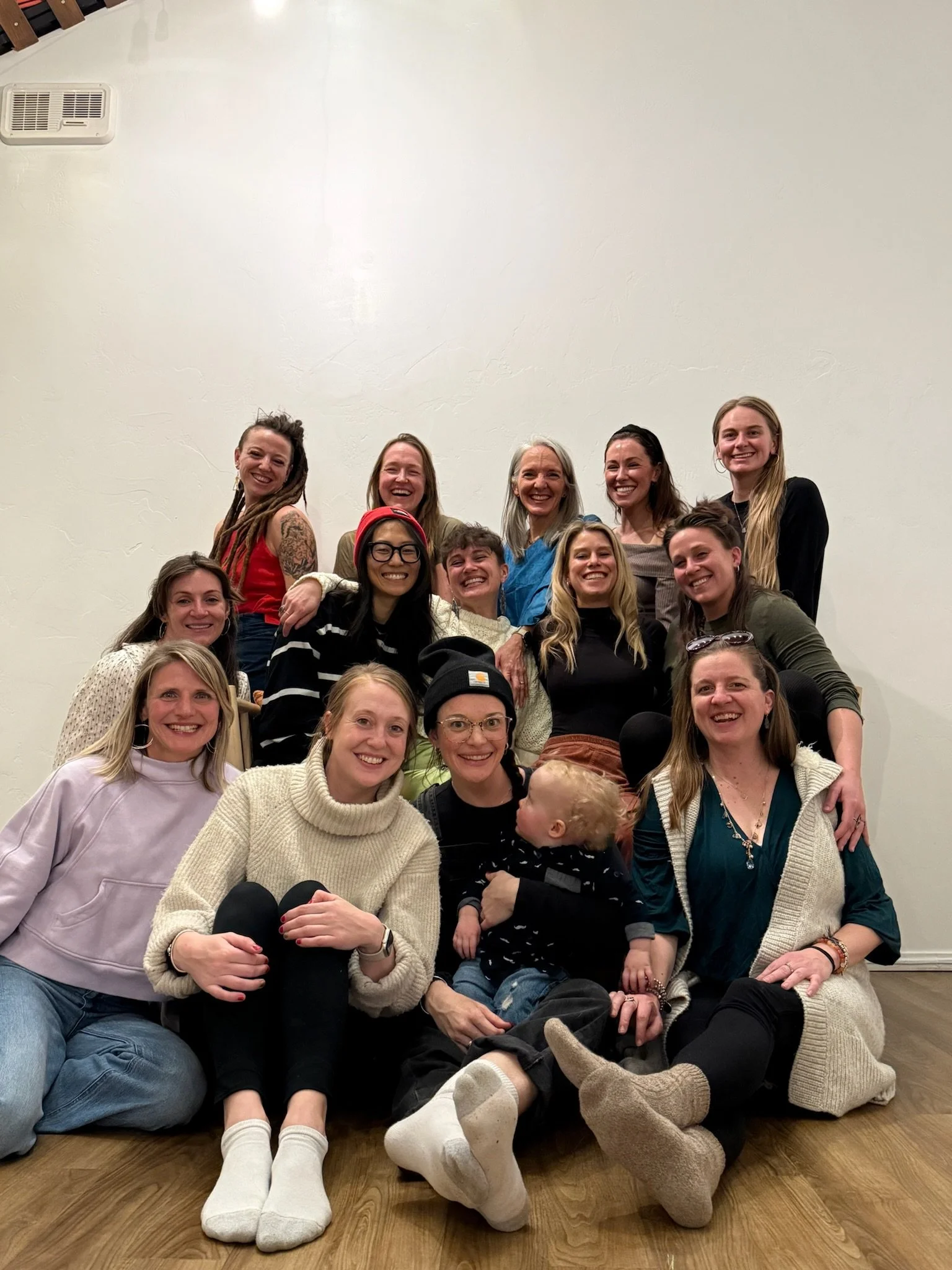 Group of smiling women and children posing together indoors with a plain white wall in the background.