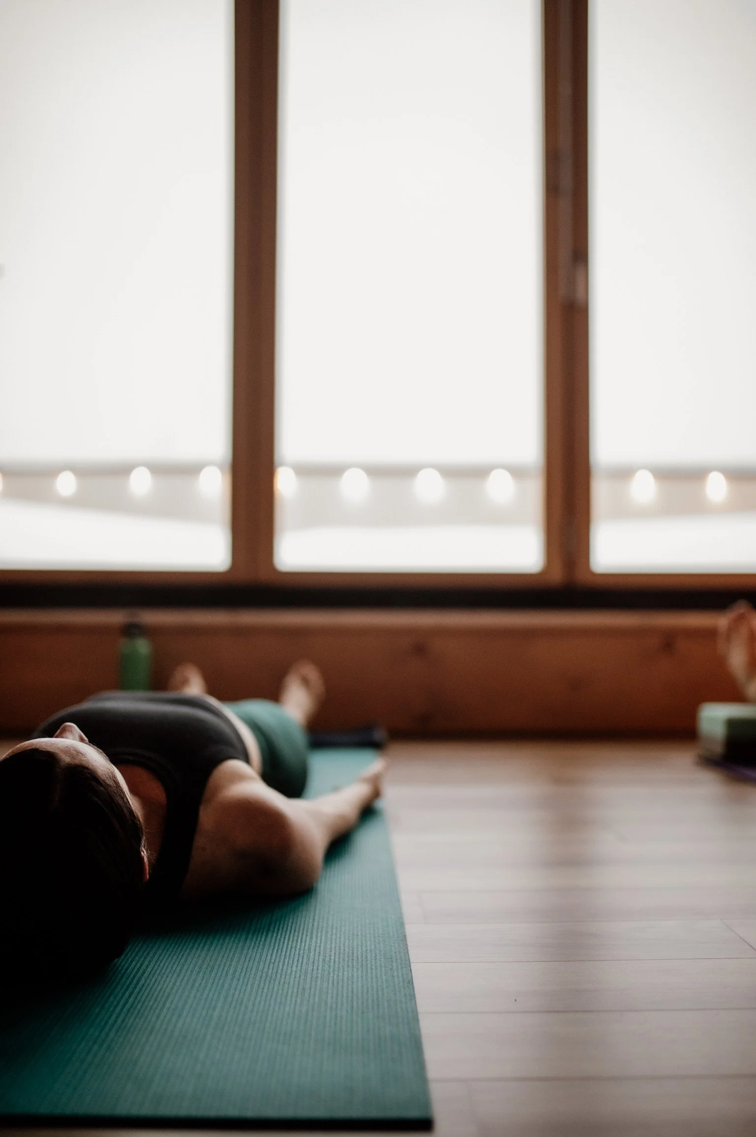 Person practicing yoga on a green mat in a room with wooden floors and large windows.