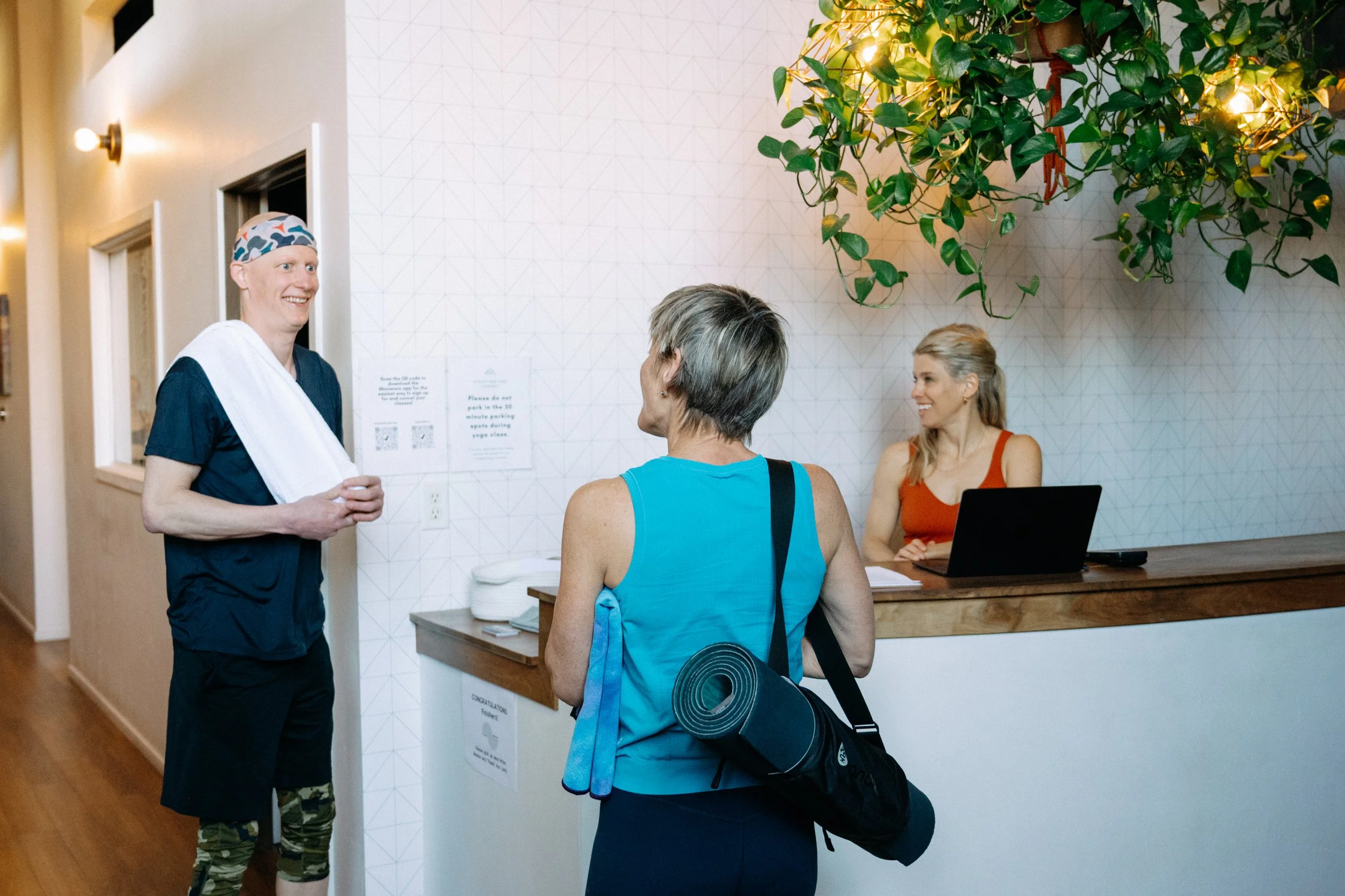 A fitness instructor smiling and talking to a woman with a yoga mat and bag, while another woman with gray hair interacts at a reception desk in a health or fitness center.