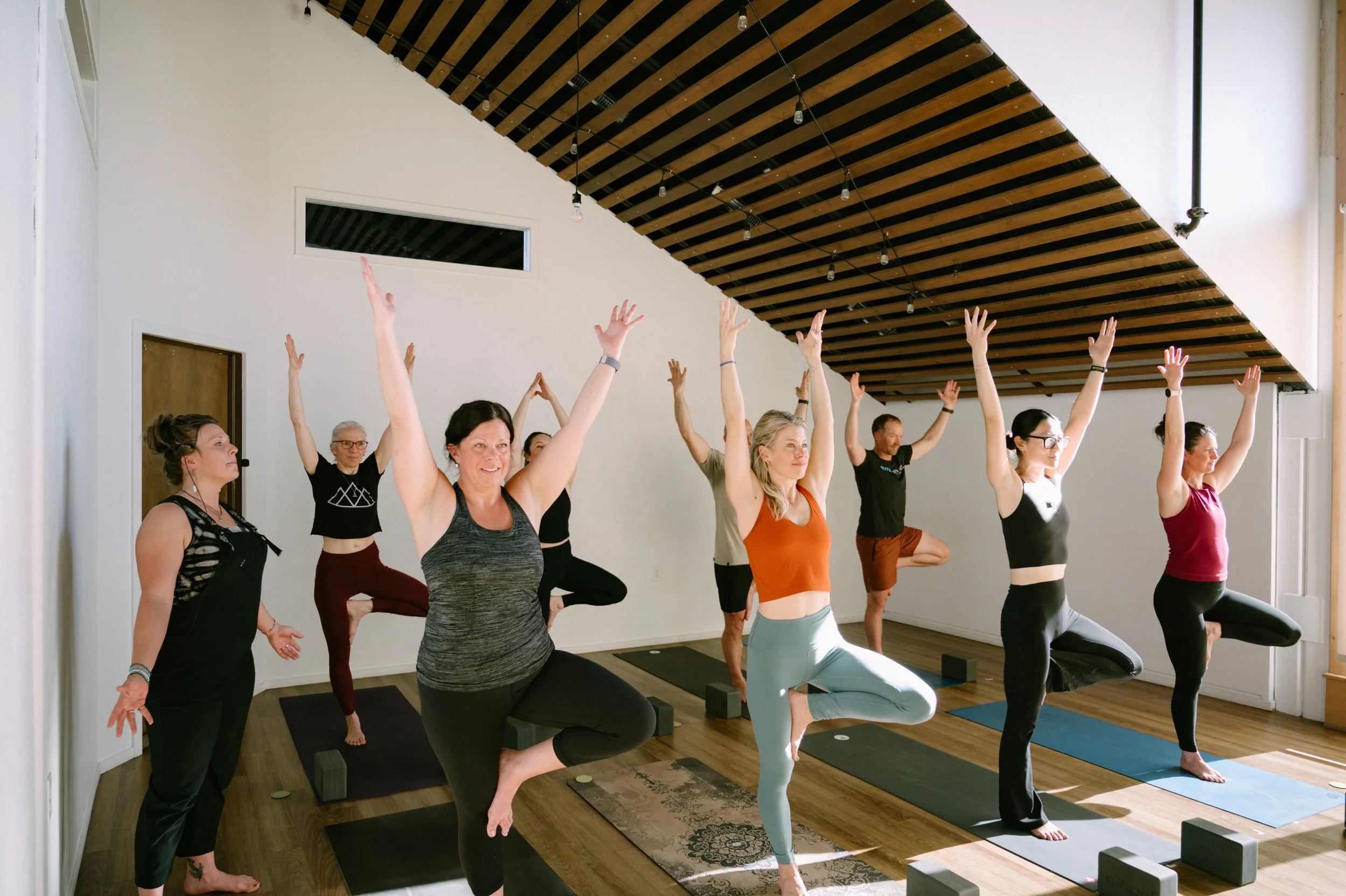 A group of people practicing yoga in a studio with wooden floors and a ceiling with wooden beams. They are standing on yoga mats, balancing on one leg with the other foot placed on their inner thigh, and raising their arms overhead.