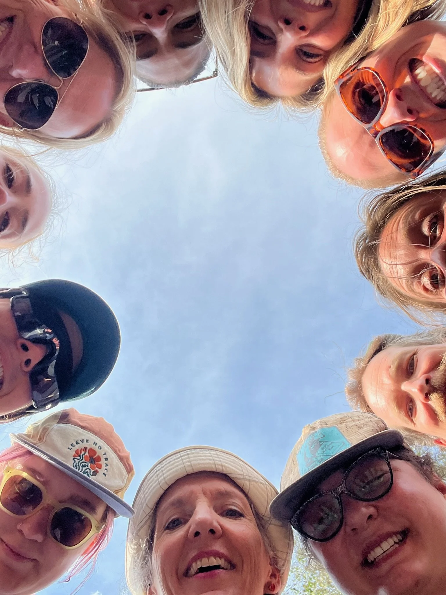 Group of people looking down at camera with smiling faces, outdoors under a blue sky, some wearing sunglasses and hats.
