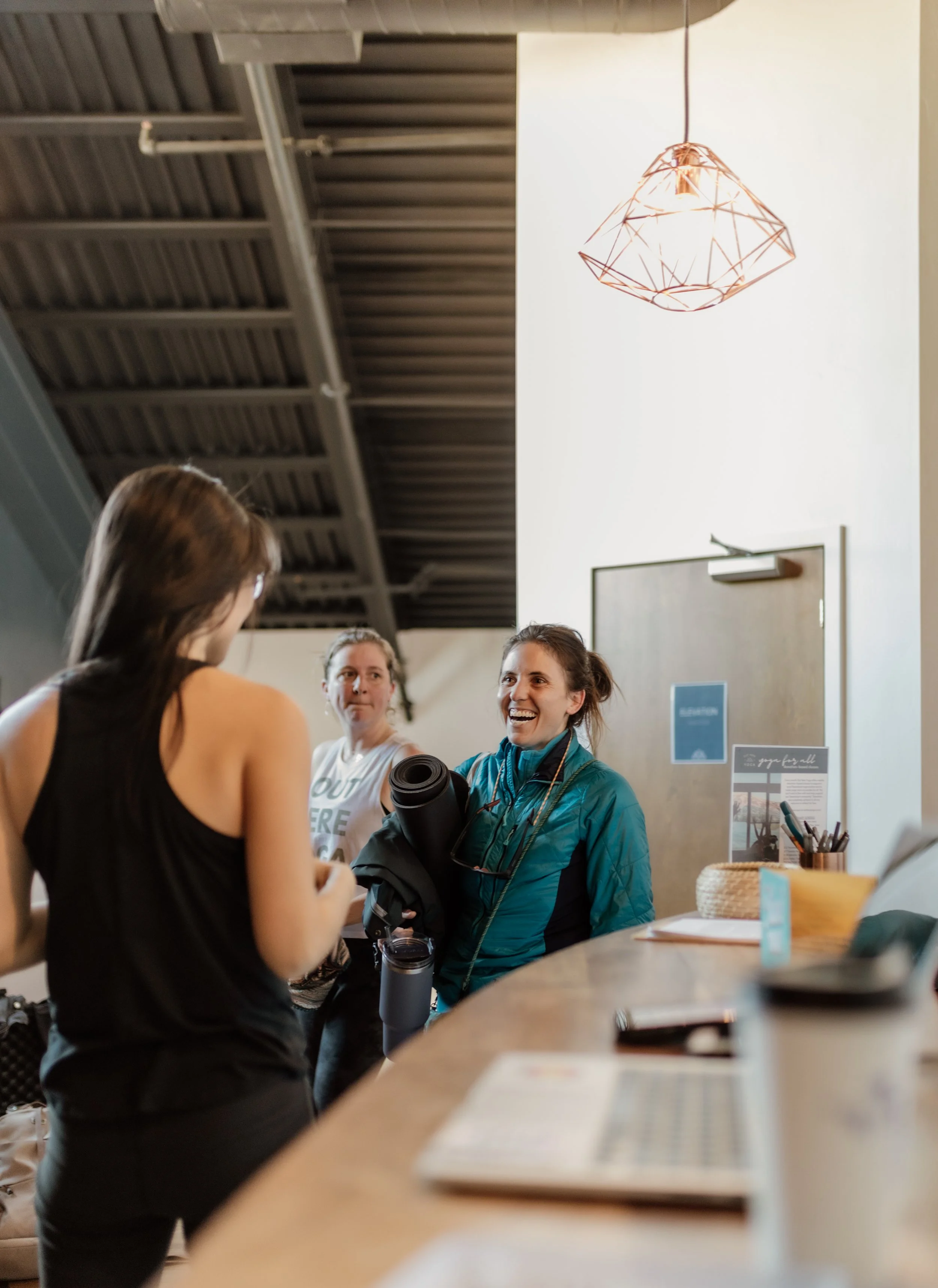Indoor scene with two women engaged in conversation, one holding a yoga mat, in a space with a high ceiling and a copper geometric pendant light.