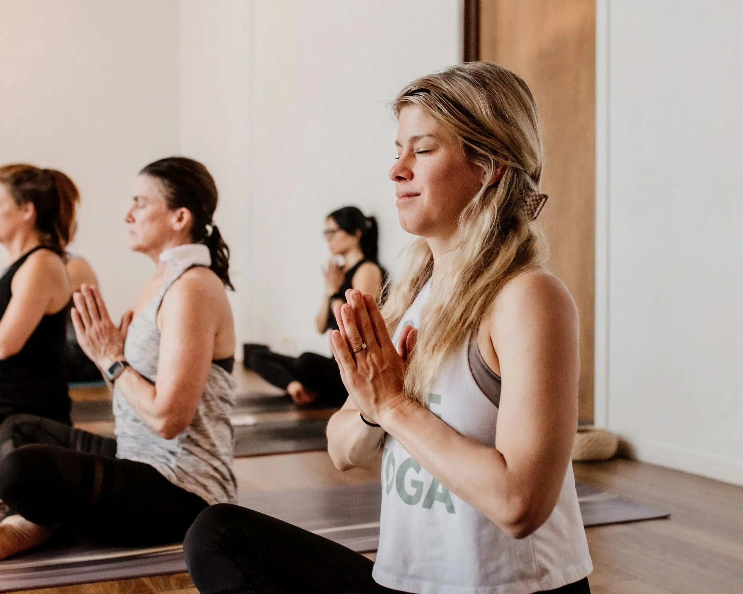 A group of women sitting on yoga mats meditating with eyes closed and hands in prayer position in a yoga class.