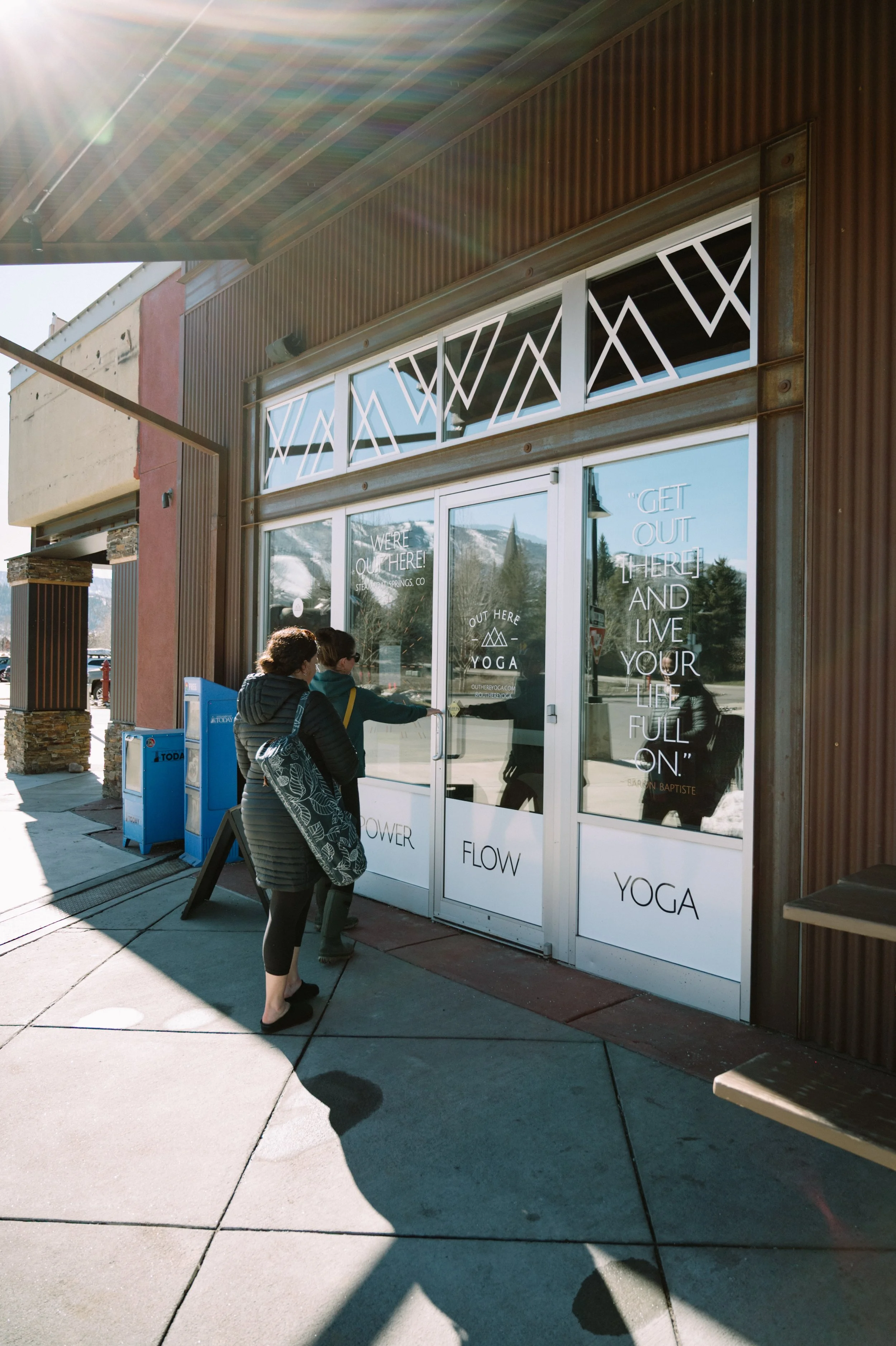 Two women are standing outside a yoga studio. The doors display inspirational quotes about living life fully. The building has a brown, textured exterior, and a sidewalk in the foreground. The weather is sunny, casting shadows on the ground.