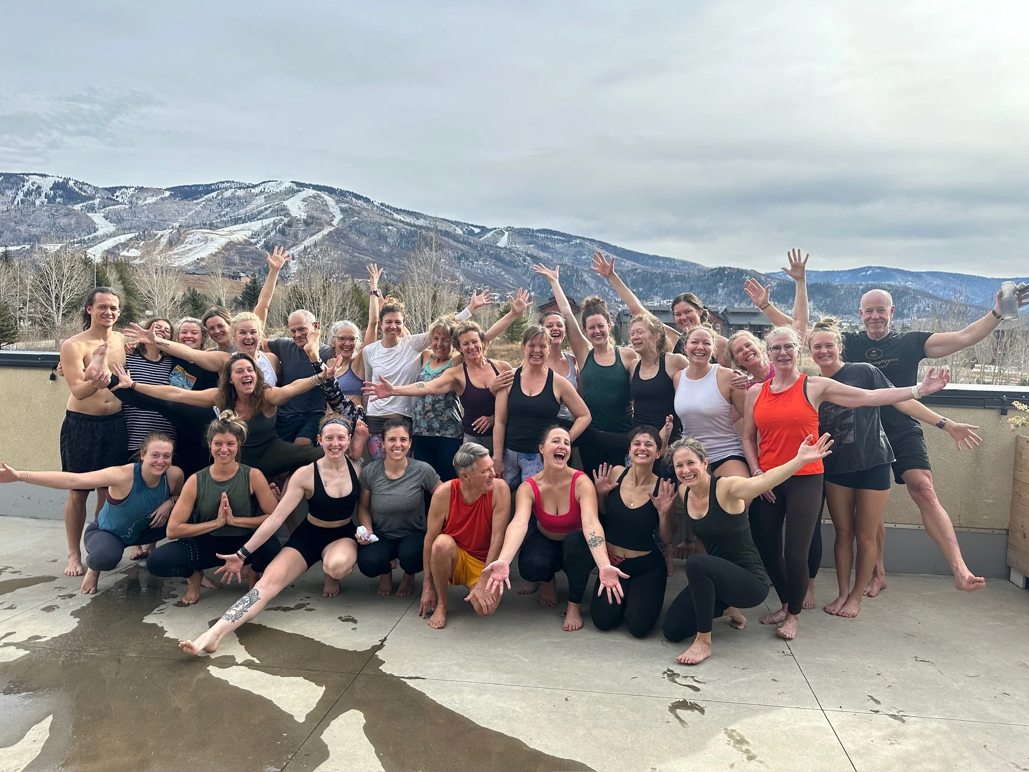 A large group of people standing on an outdoor patio with mountains and ski slopes in the background. They are smiling and making various poses, wearing casual and athletic clothing, with some barefoot.