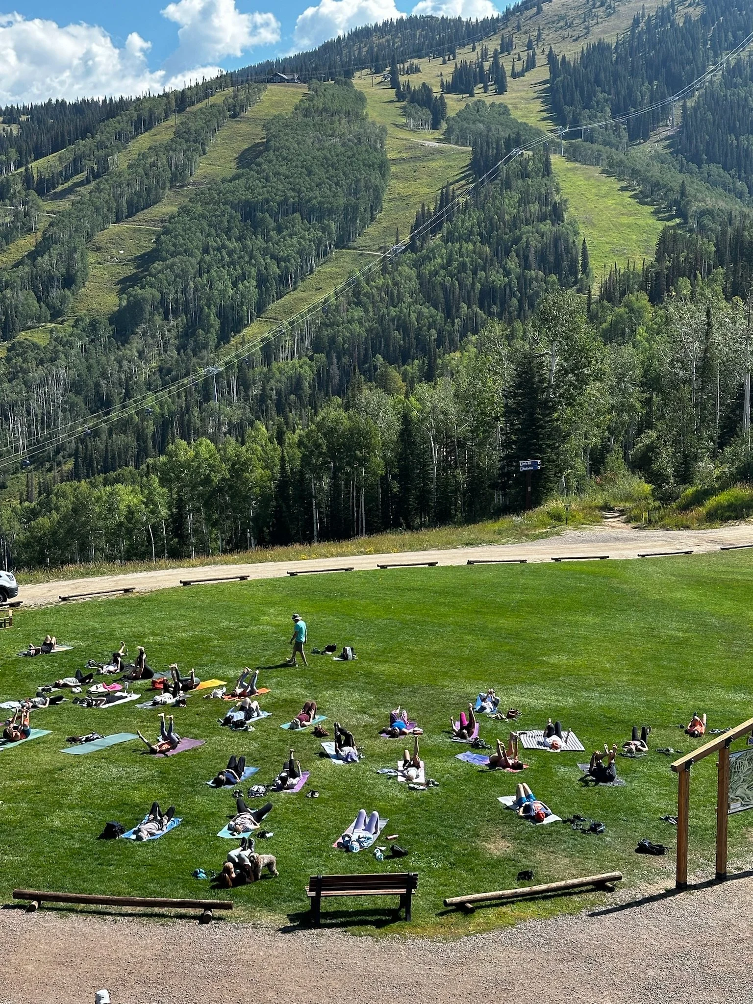 A group of people practicing yoga outdoors on a grassy hill with mountains and trees in the background, under a partly cloudy sky.