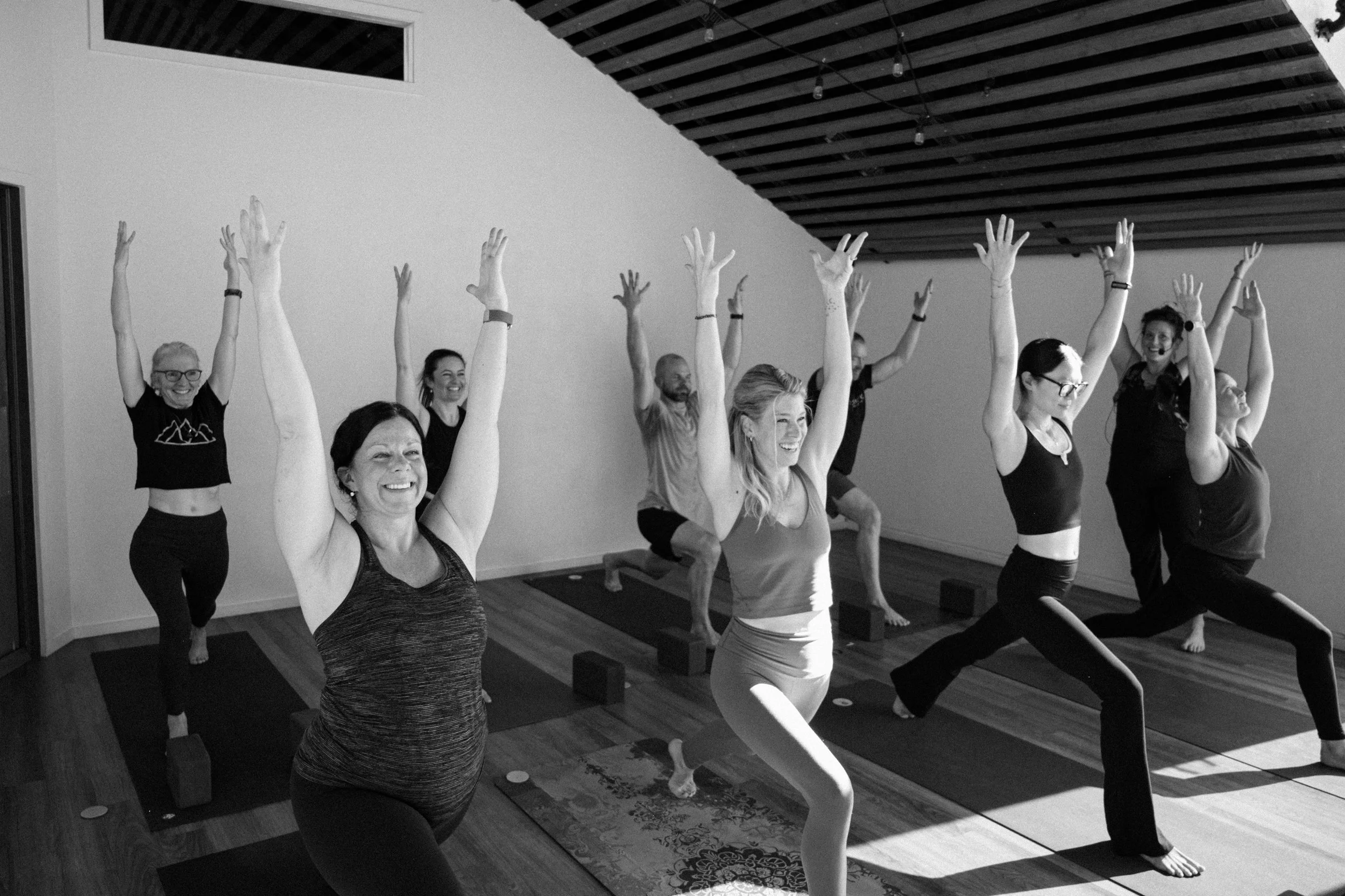 A group of diverse women and men participating in a yoga class indoors, standing on yoga mats with arms raised above their heads, smiling and appearing focused, in a well-lit room with wooden floors and a slanted ceiling with string lights.