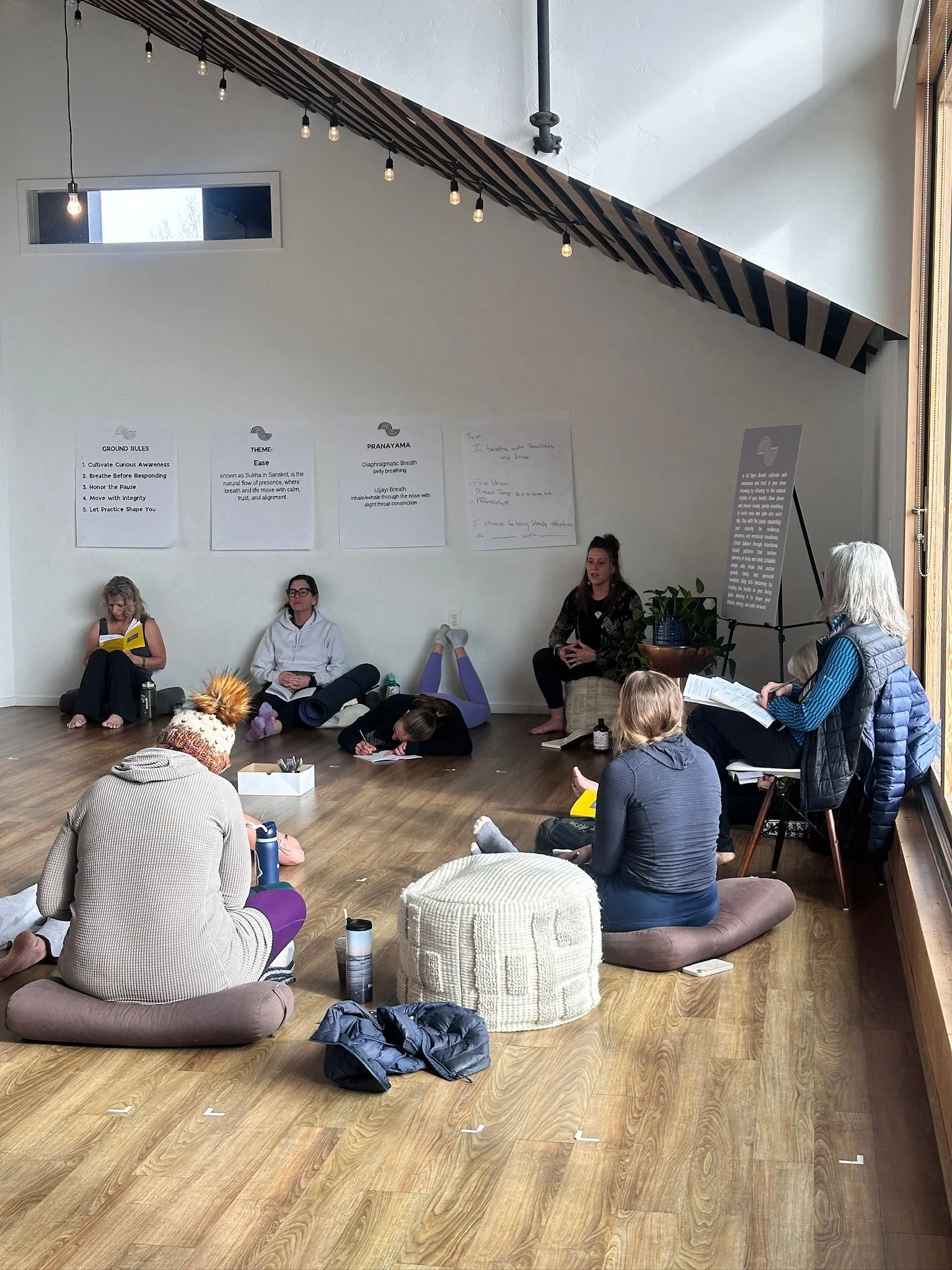 Group of people participating in a workshop or meditation session in a cozy room with wooden flooring, sitting on cushions and mats, with some taking notes or reading, and a few with meditation or yoga props around them.