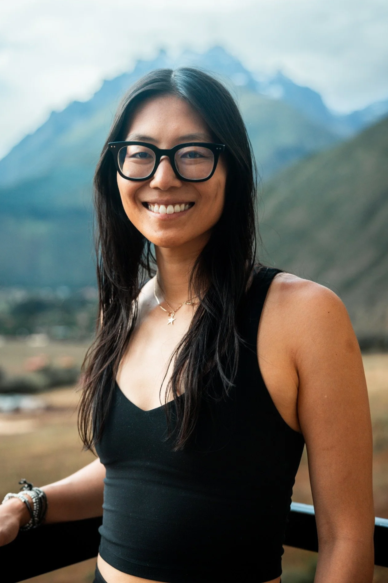 A smiling woman with black hair and glasses outdoors with mountains in the background, wearing a black sleeveless top and layered necklaces.