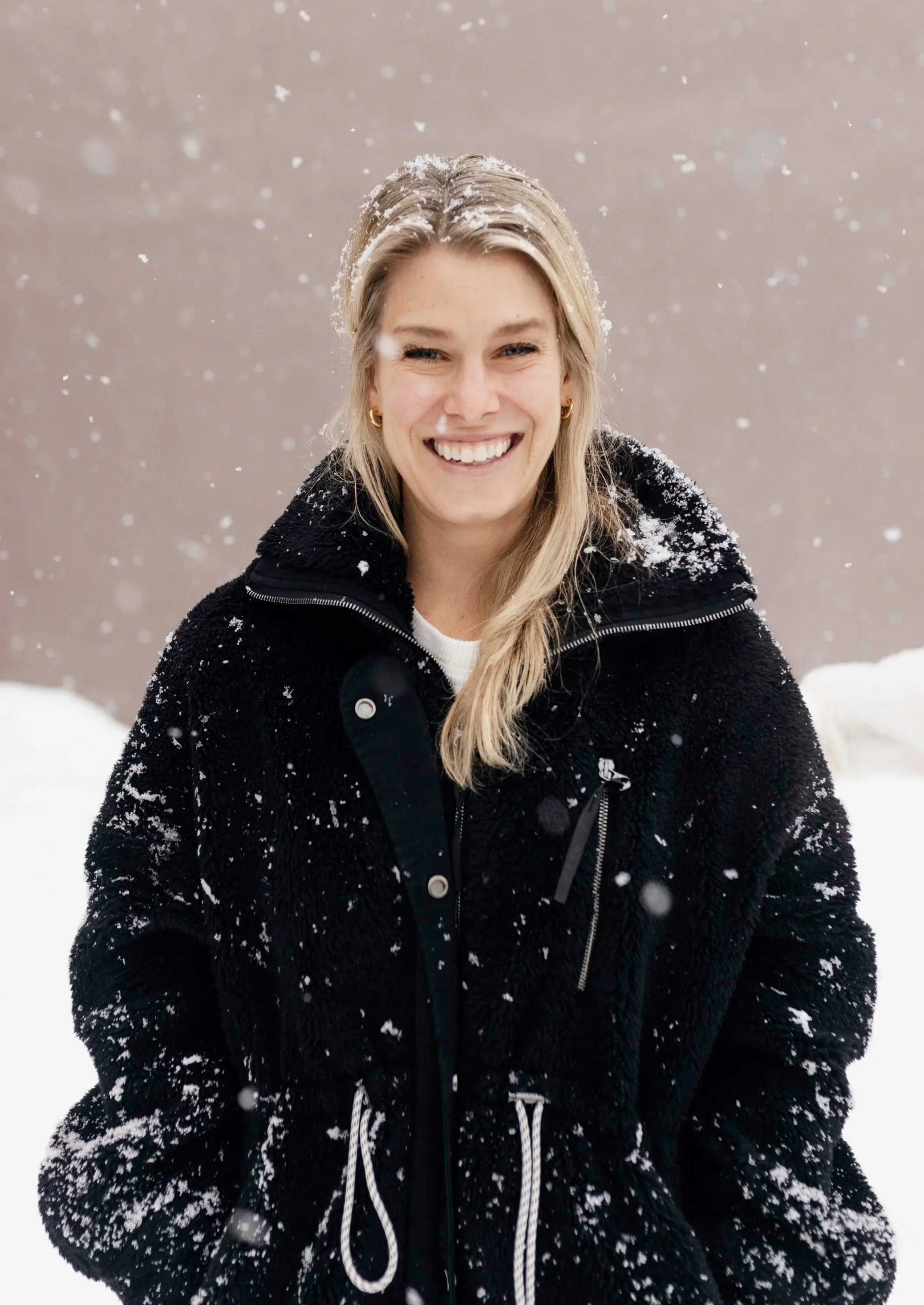 A smiling woman with long blonde hair and gold earrings, wearing a black jacket with snow on it, standing outdoors in a snowy landscape with snow falling around her.