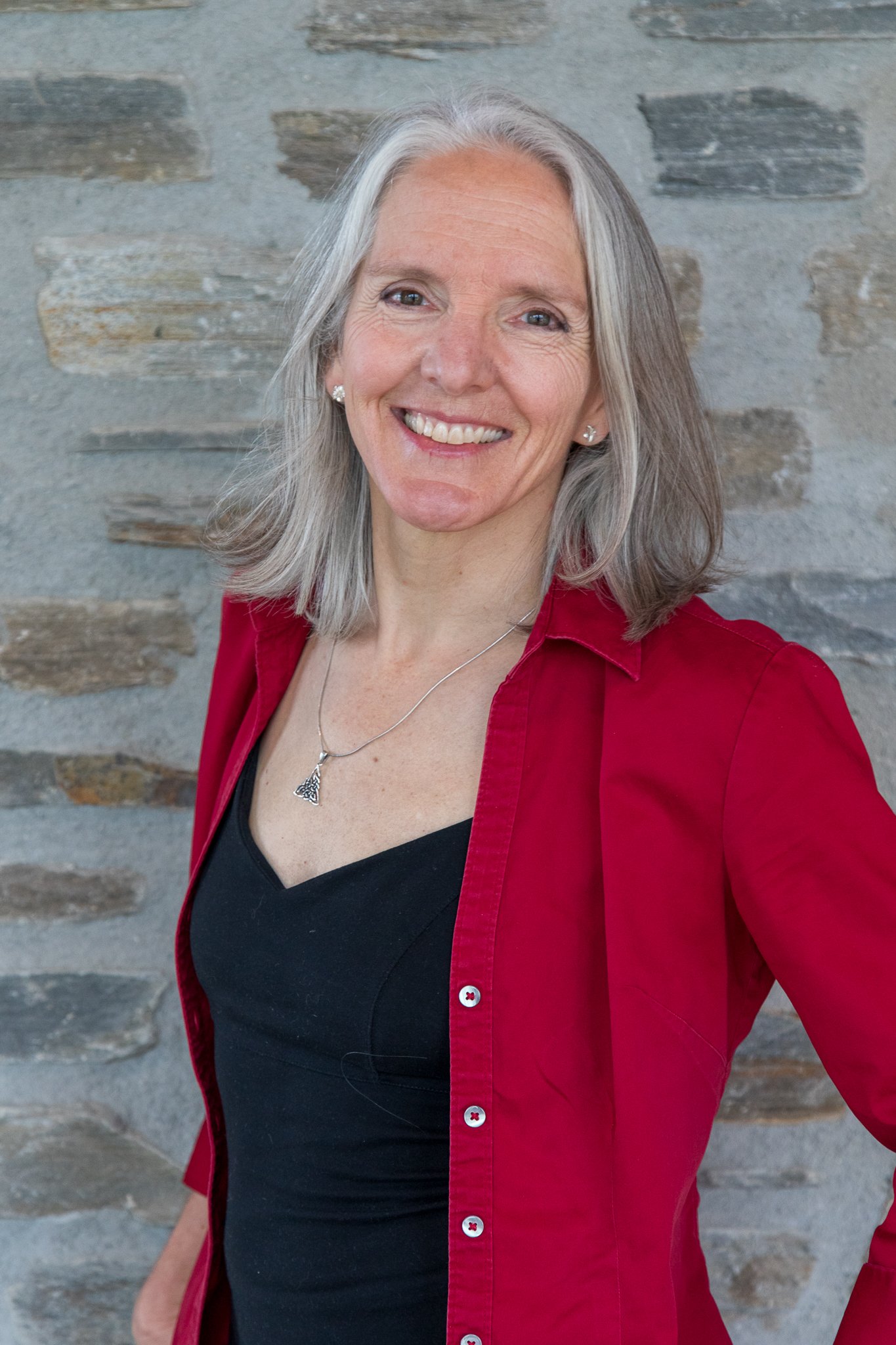 A smiling middle-aged woman with gray hair wearing a black top and a red button-up shirt, standing in front of a stone wall.