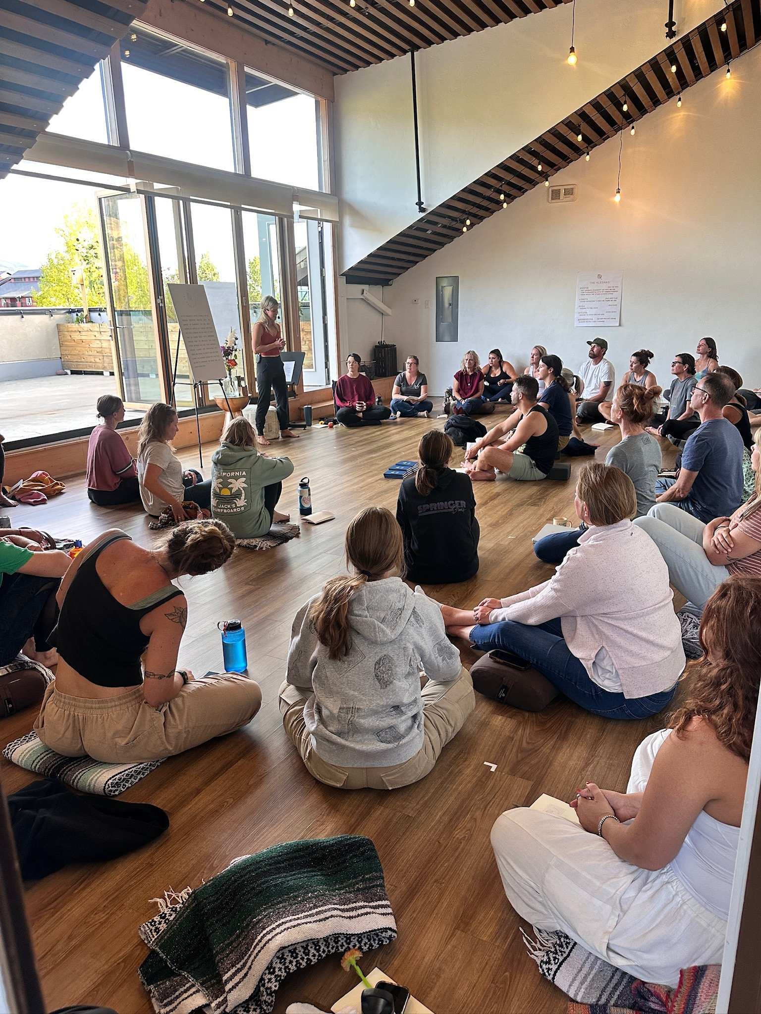 Group of people sitting on the floor in a room with high ceilings, large windows, and hanging string lights, attending a presentation or workshop with a woman speaking at the front.