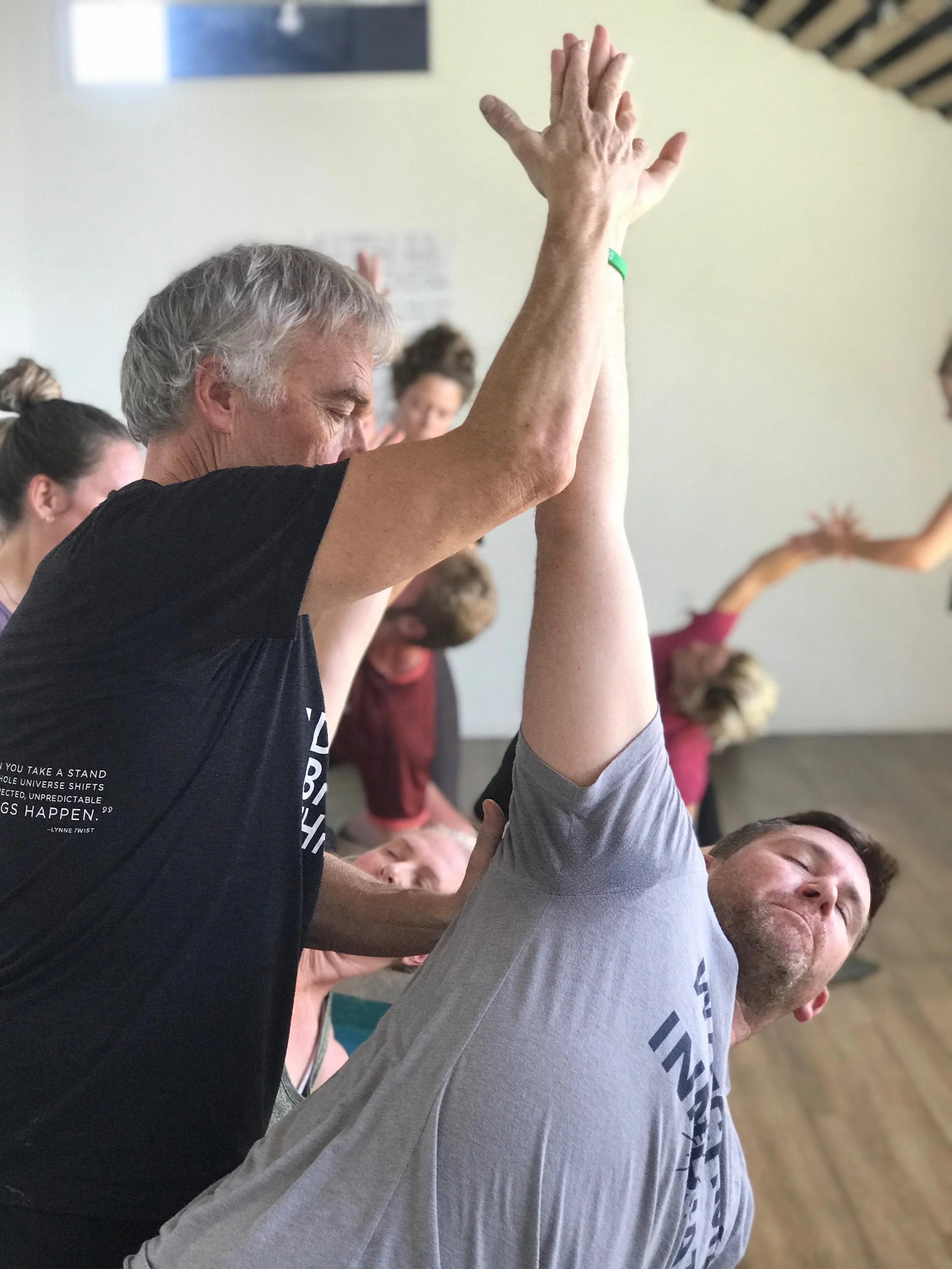 People practicing yoga in a studio, with a man assisting a student in a yoga pose by pressing their hands together.