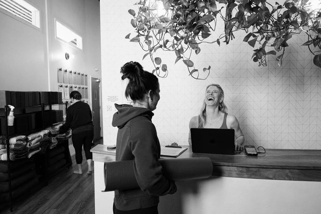 A woman greets a guest at a minimalist hotel reception desk, laughing and engaging with her, as another staff member arranges linens in the background.