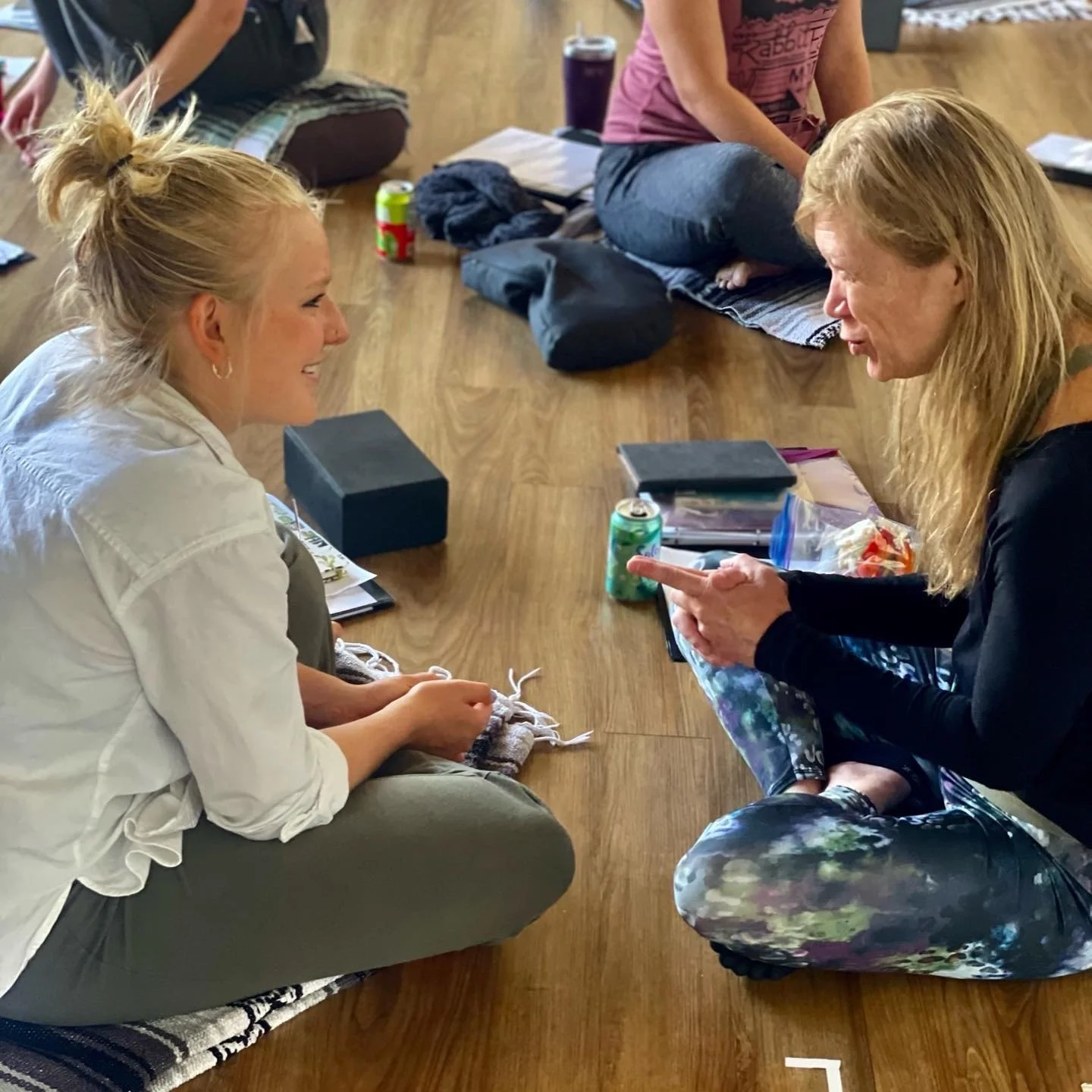A woman with long blonde hair wearing colorful leggings and a black top, seated on the floor, talking to a smiling woman with her hair in a bun, wearing a white shirt and gray pants, also seated on the floor, in a cozy indoor setting with other people in the background.