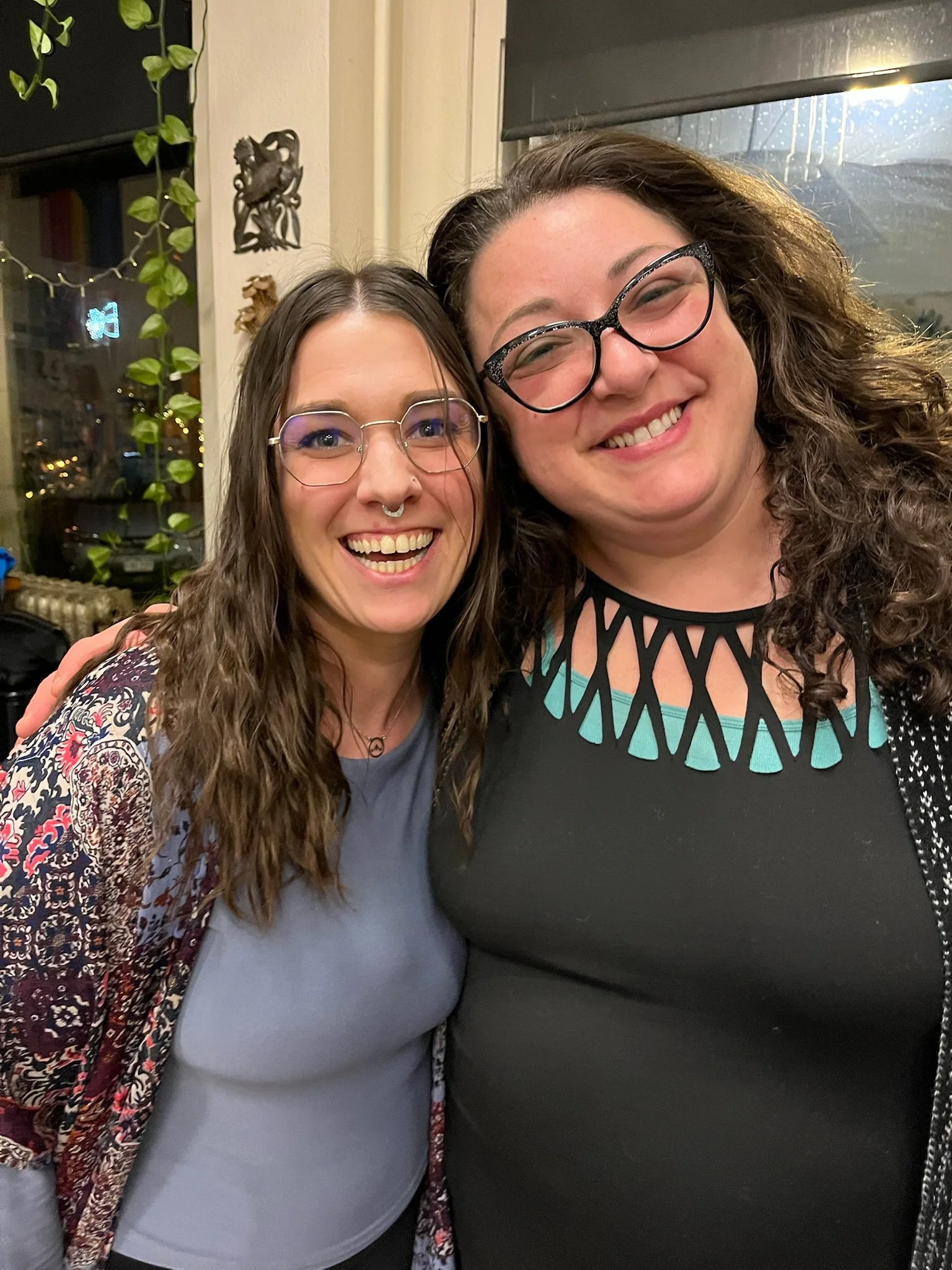 Two women smiling for a photo, one has glasses and a septum piercing, the other has curly hair and glasses.