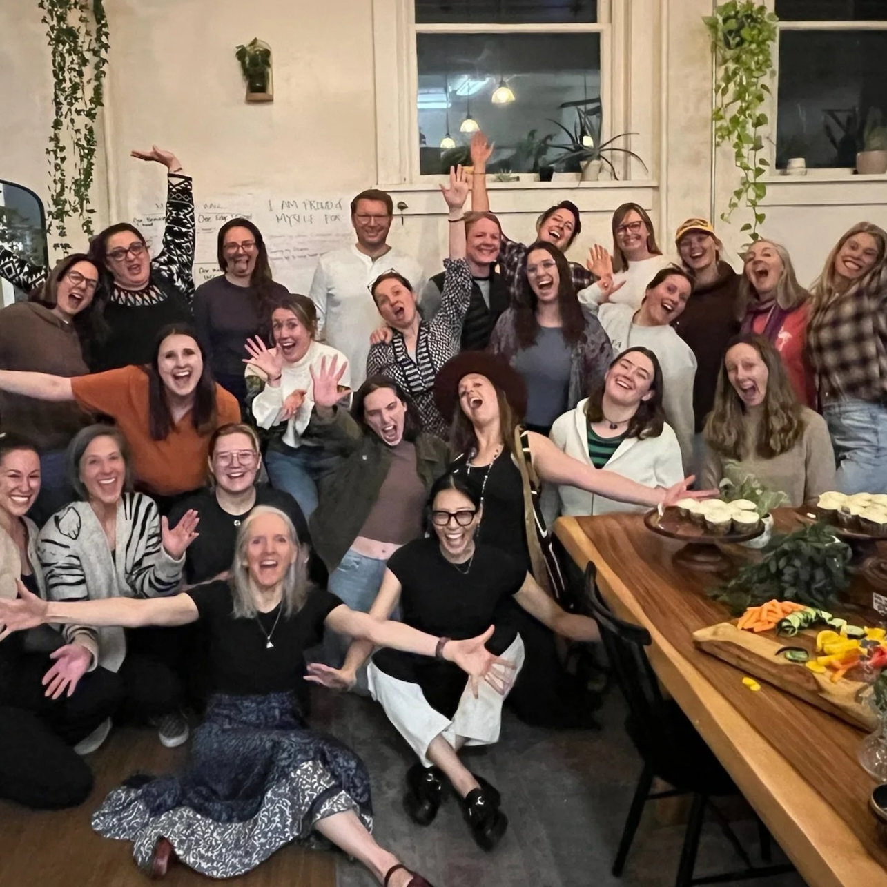 A large group of people gathered indoors at a celebration, smiling and laughing with arms raised and outstretched, with a table of food on the right side of the image.