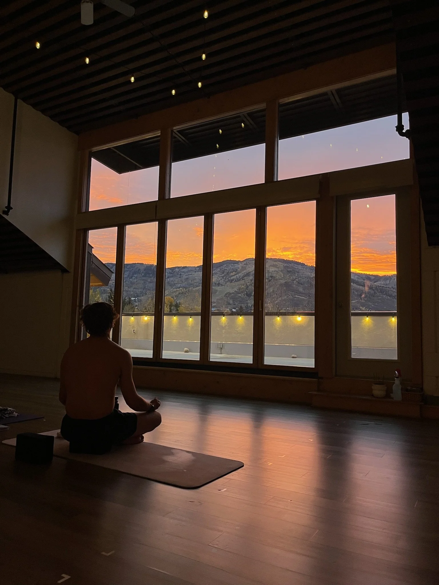 A person sits cross-legged on a yoga mat inside a room during sunset, with large windows showing mountains and a colorful sky outside. The room has string lights hanging from the ceiling.