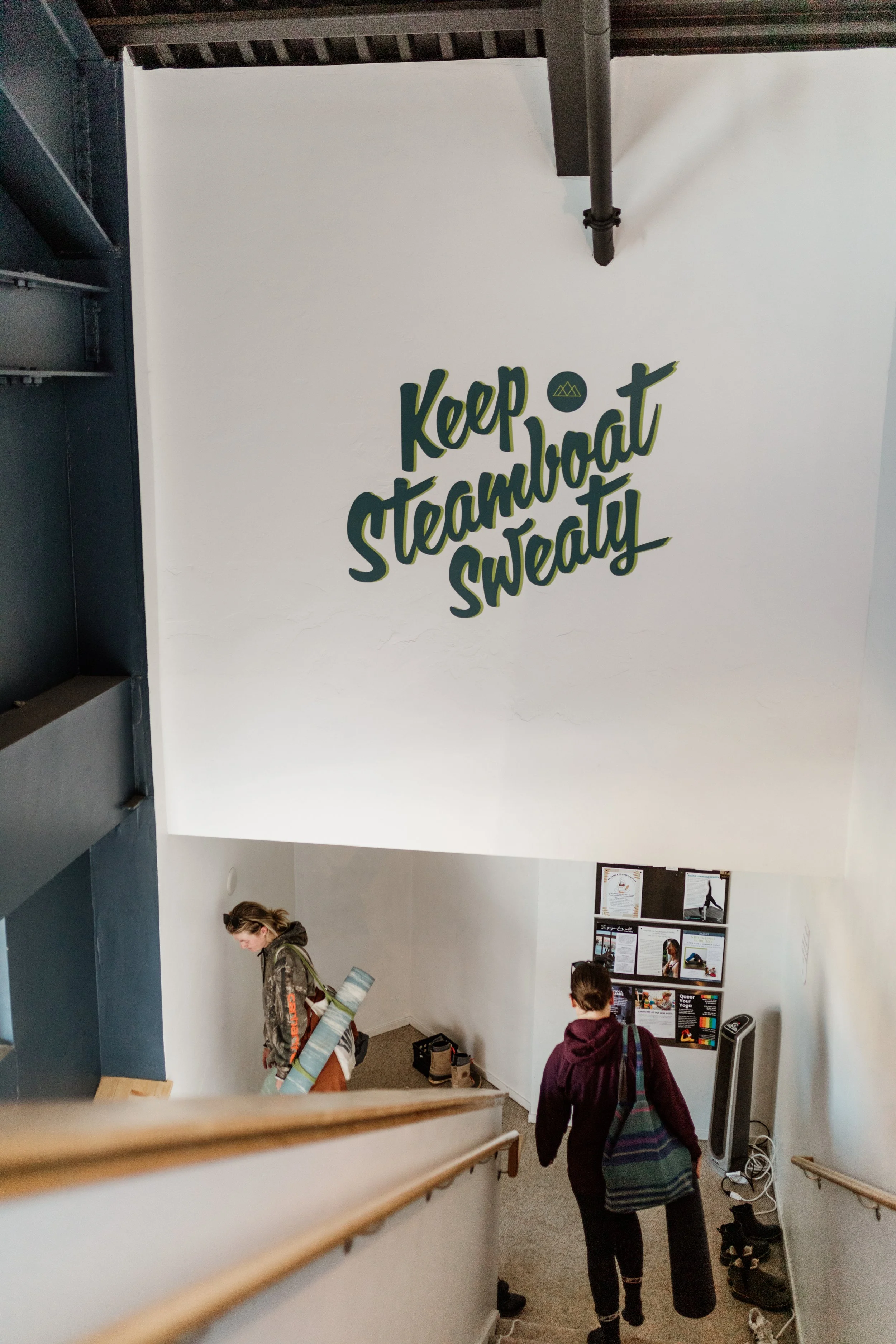 Inside a building with a large hanging sign that reads, 'Keep Steamboat Sweaty.' The staircase is visible, and a woman with a yoga mat and backpack is descending. Two people are at the bottom, one with a backpack.