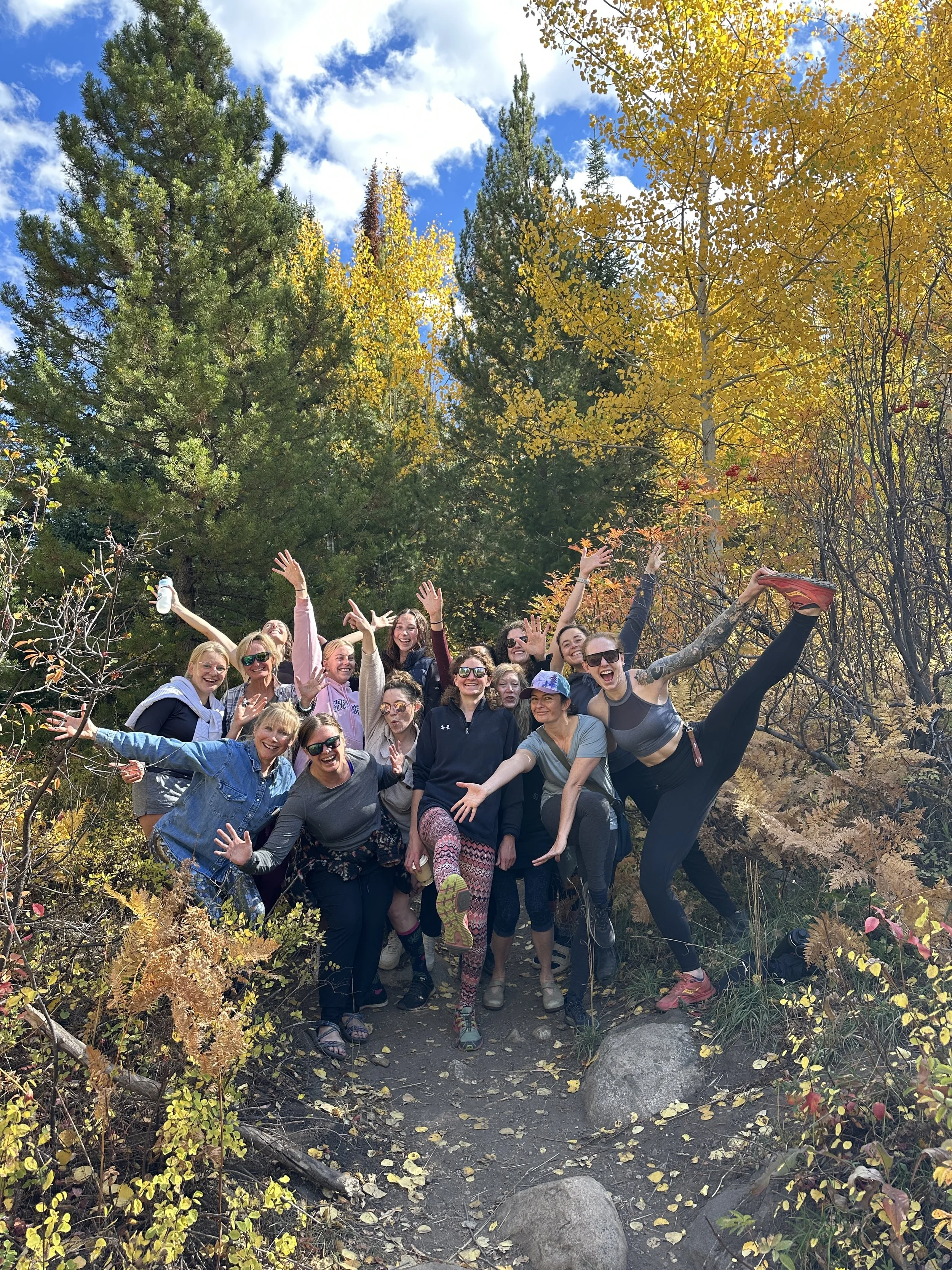 Group of smiling women on a fall hike in forest with yellow and orange leaves, blue sky, and clouds.