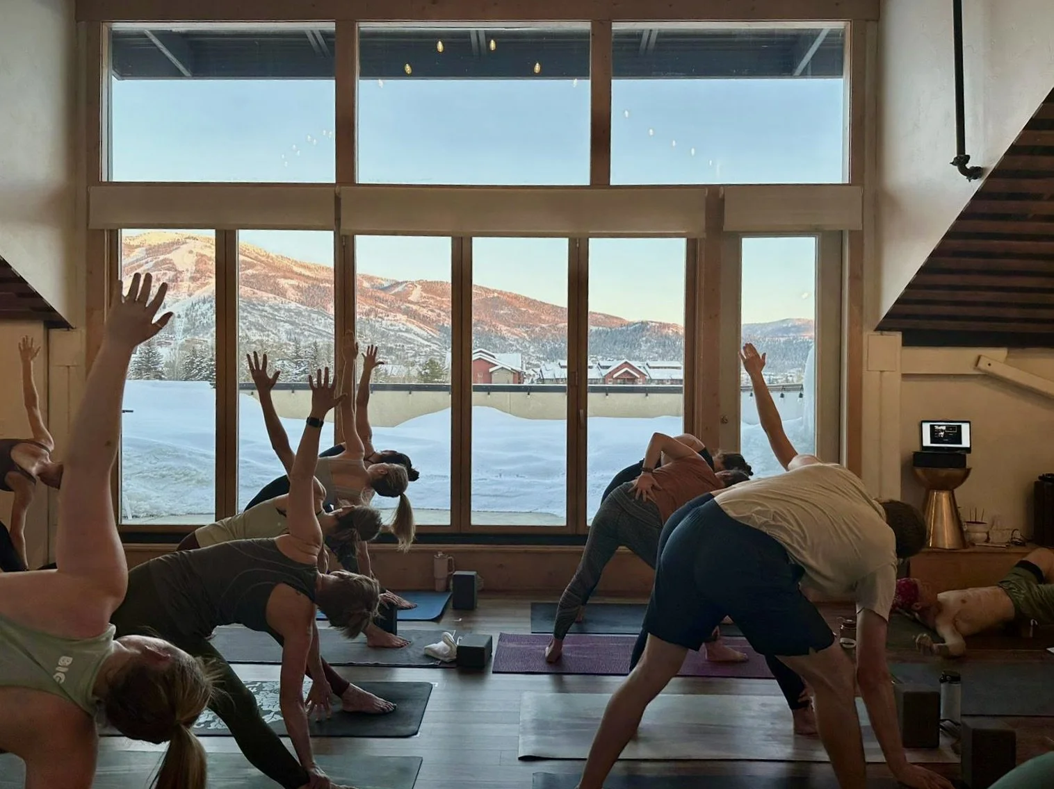 People participating in a yoga class inside a wooden-paneled room with large windows overlooking a snowy landscape and mountains.