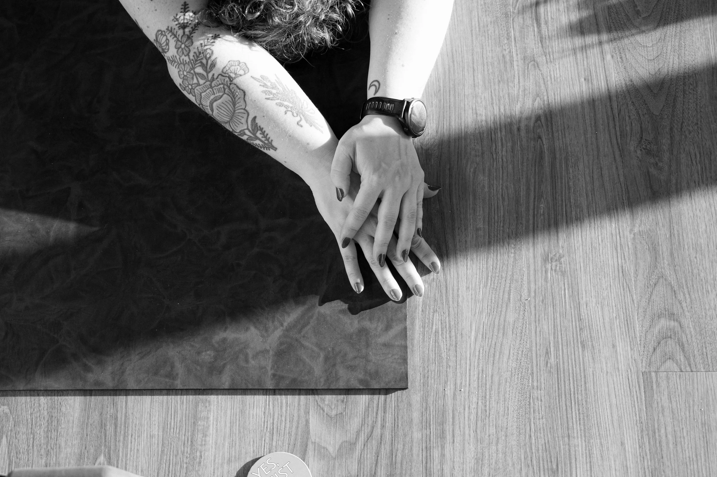 A person's hands with nail polish resting on a wooden surface. The person has tattoos on their forearm and a wristwatch. There is a piece of paper on the surface with text that reads 'YES, PLEASE!'
