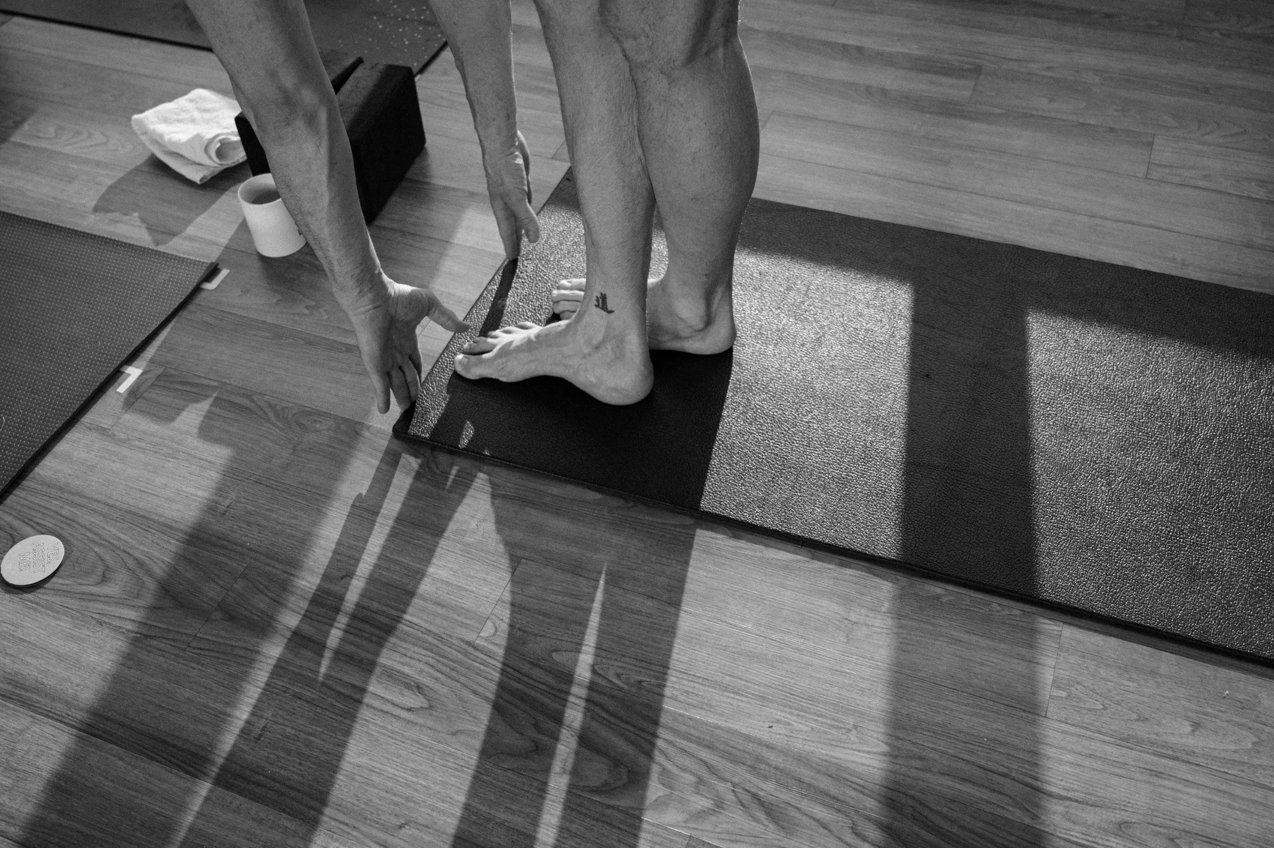 A person is stretching on a yoga mat on a wooden floor, reaching for their feet in a forward fold with feet crossed, surrounded by a rolled towel, and a mug.